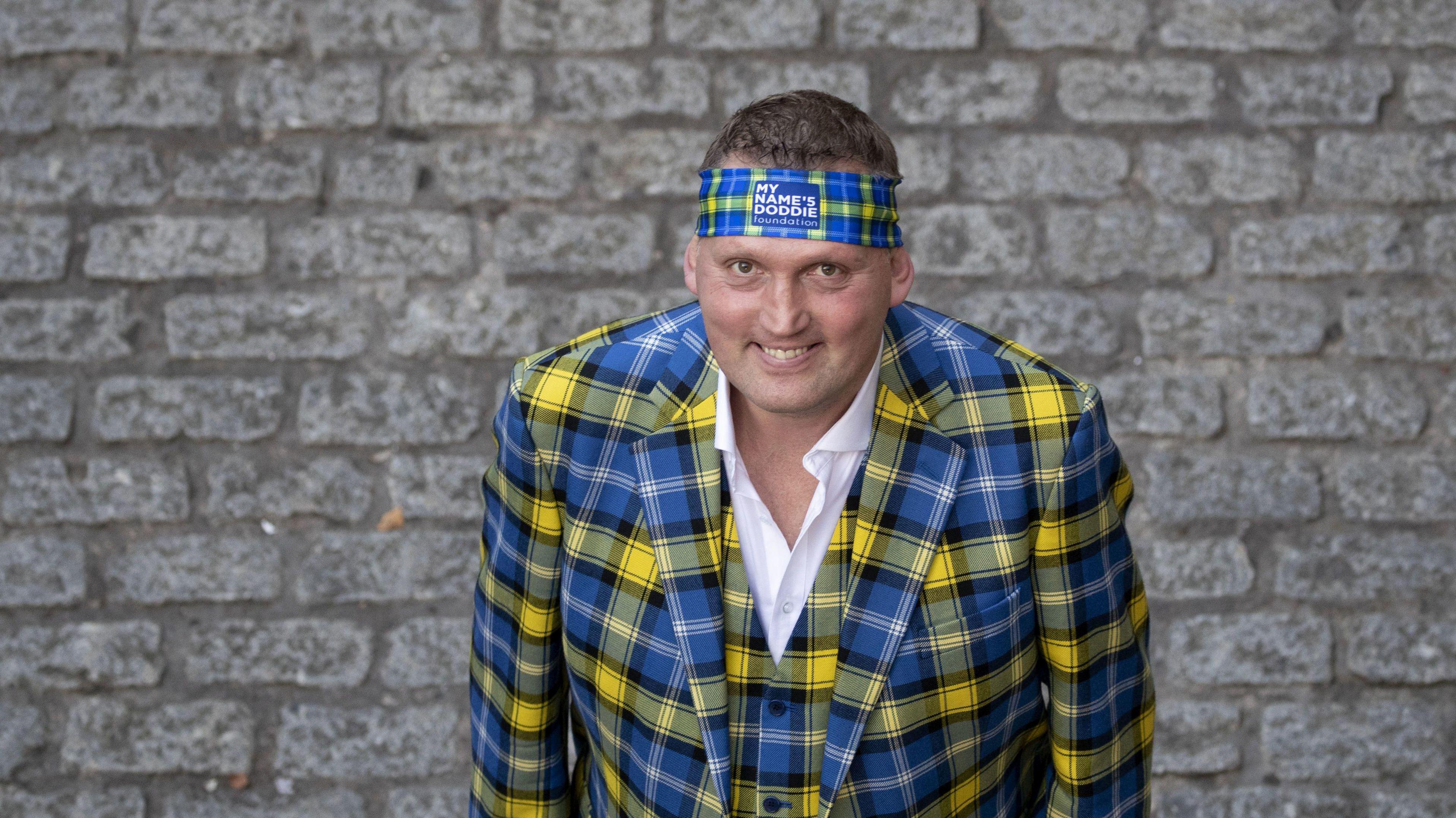 Doddie Weir wearing a blue and yellow tartan suit and a matching headband. He has short brown hair.