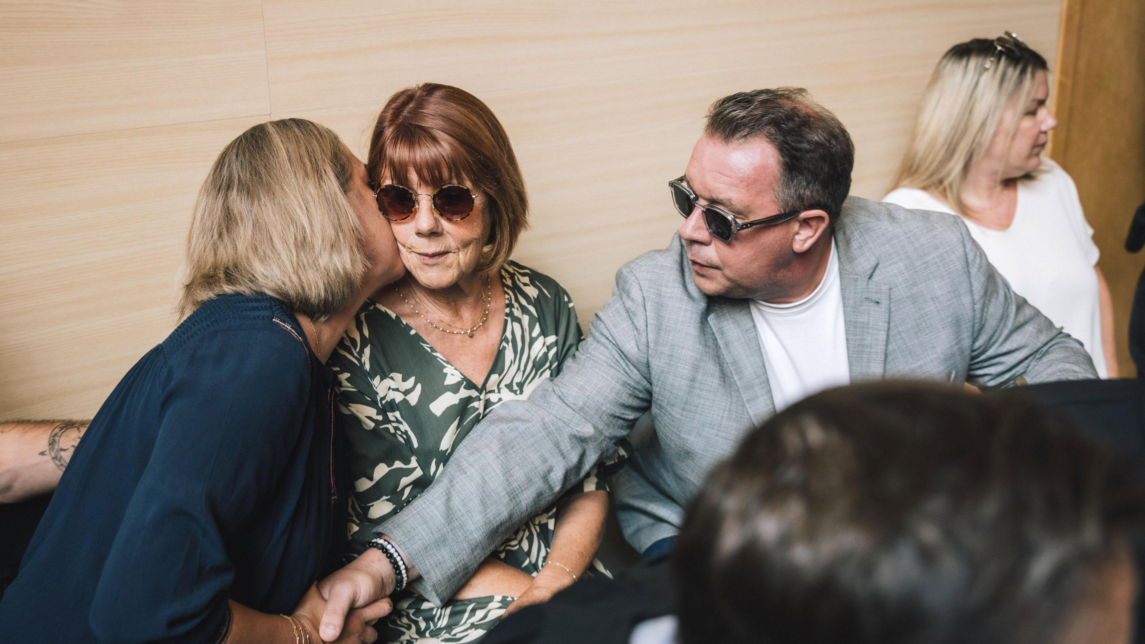 Gisèle Pelicot sitting in Avignon Courthouse, wearing round tortoiseshell sunglasses, as her daughter Caroline leans over to kiss her on the cheek. At the same time, David, also wearing sunglasses, reaches over to hold Caroline's hand.