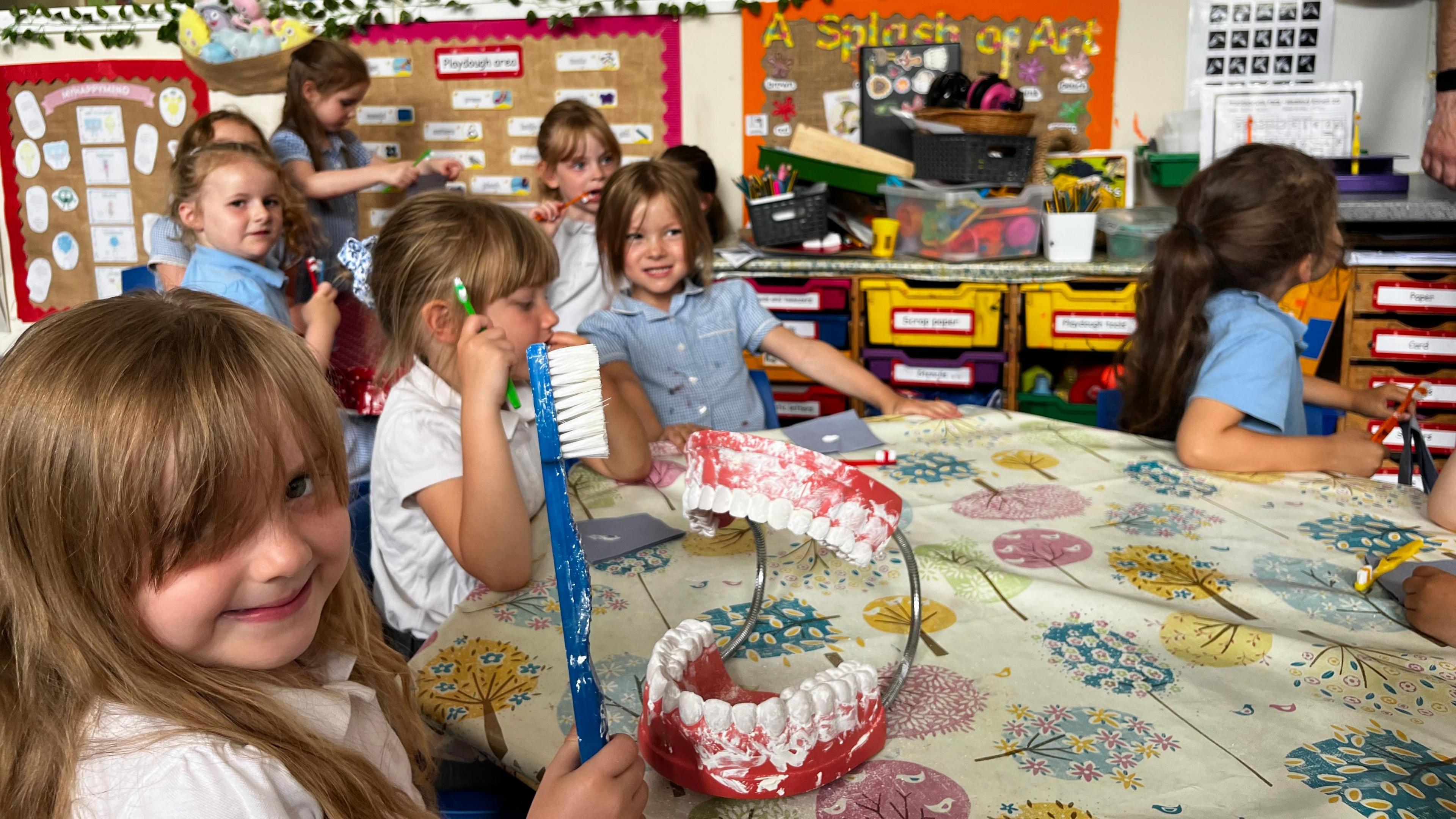 The image shows nine school girls sat in a class room brushing their teeth. One girl sat near the front is brushing an extra large fake set of teeth.