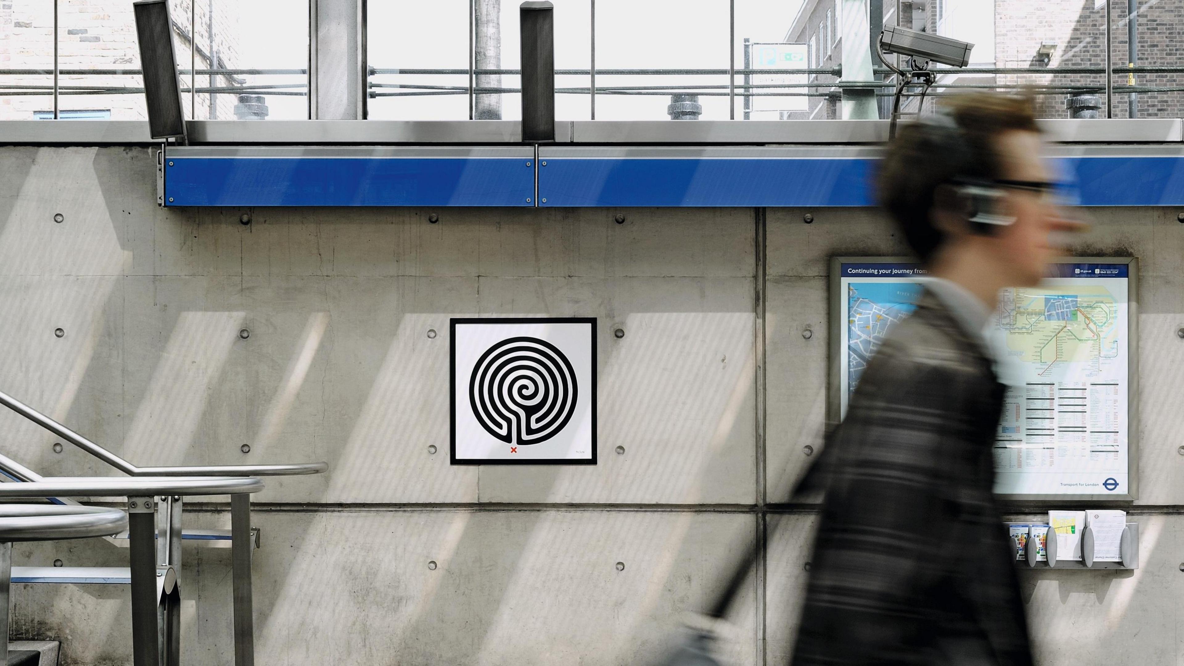 A small plaque is attached to the grey concrete wall of a modern Tube station. It has a black and white image that resembles a labyrinth on it. A person walks past, his image is blurred. 