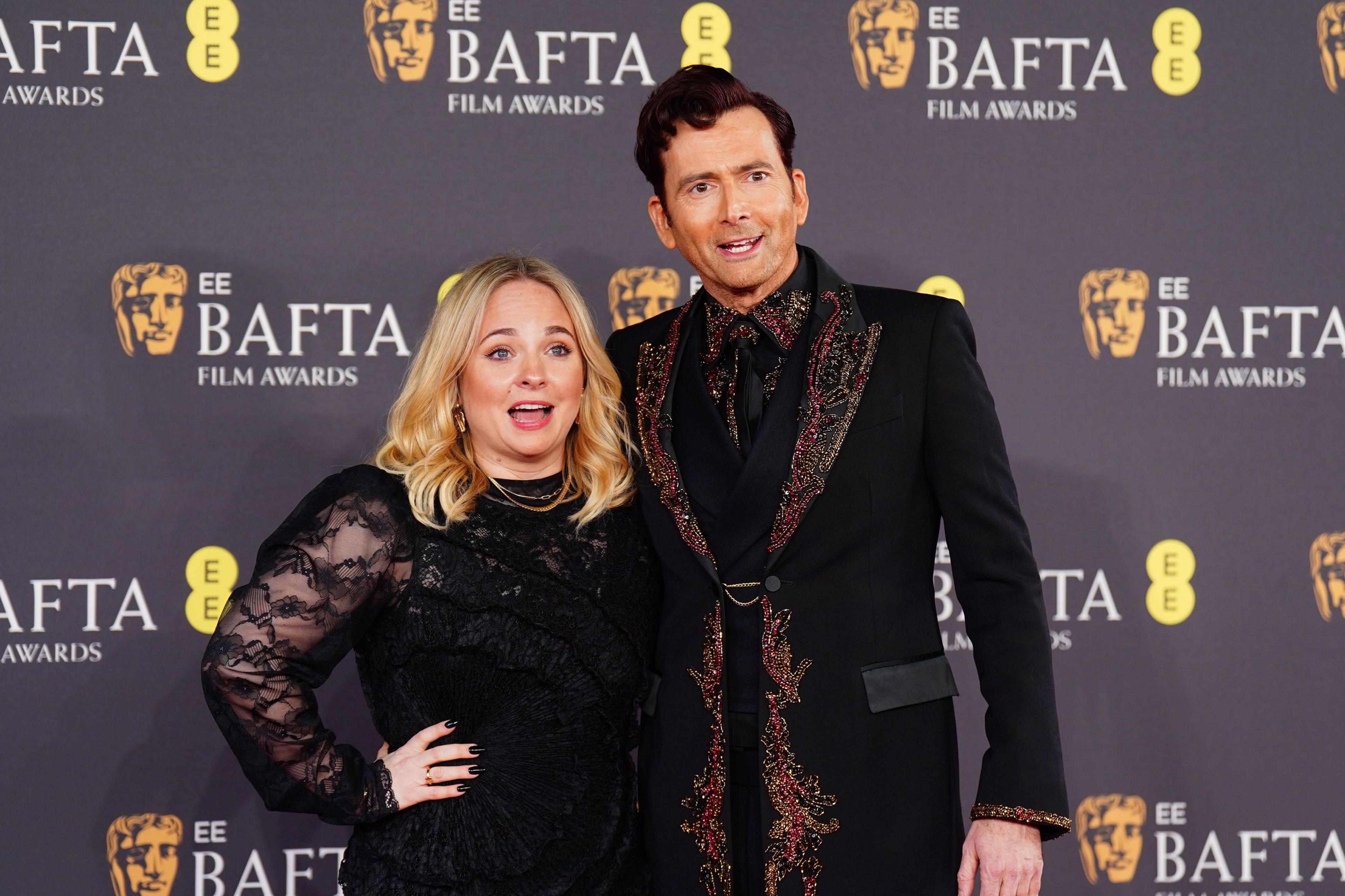 David and Georgia Tennant in black outfits on the red carpet, David's jacket and shirt have red jewels on the collars and lapels