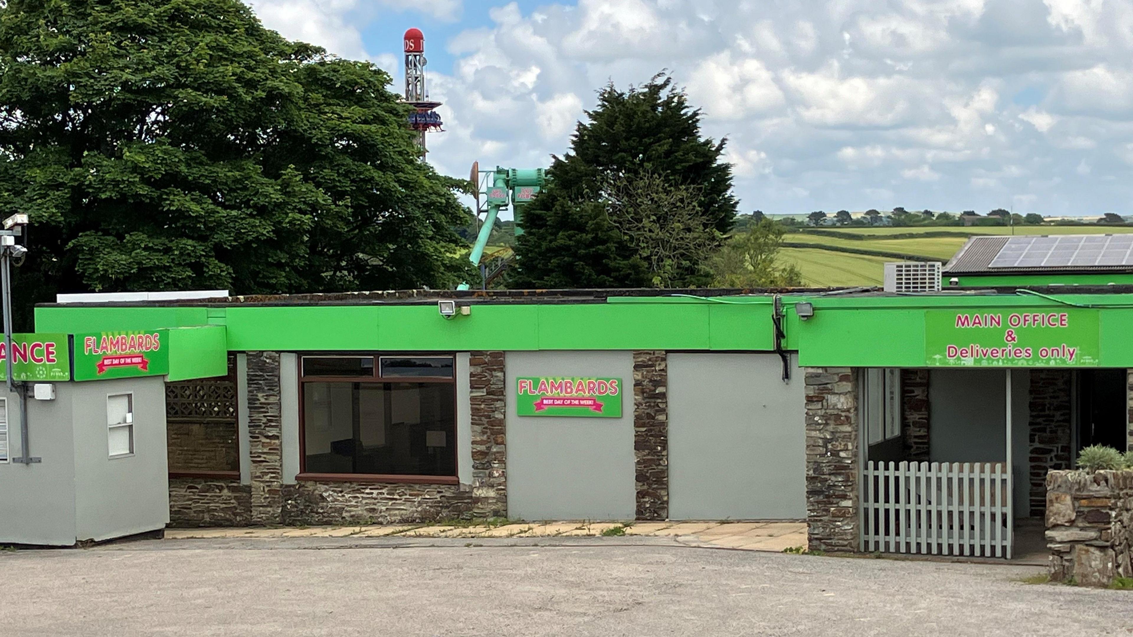 The entrance of Flambards Theme Park. A single story stone façade building with bright green signage and red letters provides a gate to the park. Rides trees and fields in the background.