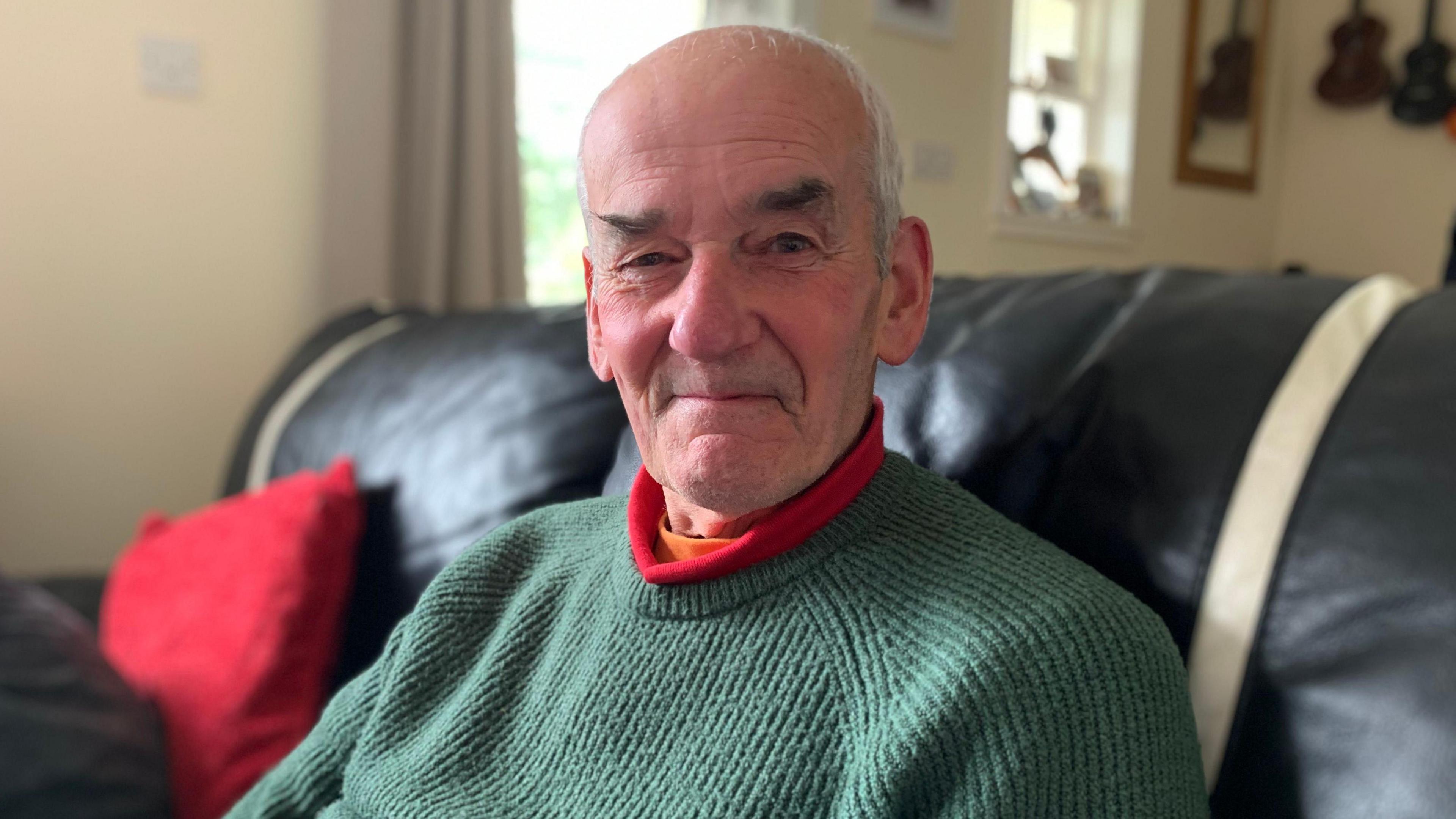 A bald headed man with grey hair and bushy eyebrows in a thick green jumper with a red collar popping out from underneath. He is sitting in a living room with a red cushion on a black sofa and some musical instruments are visible hanging from the wall.