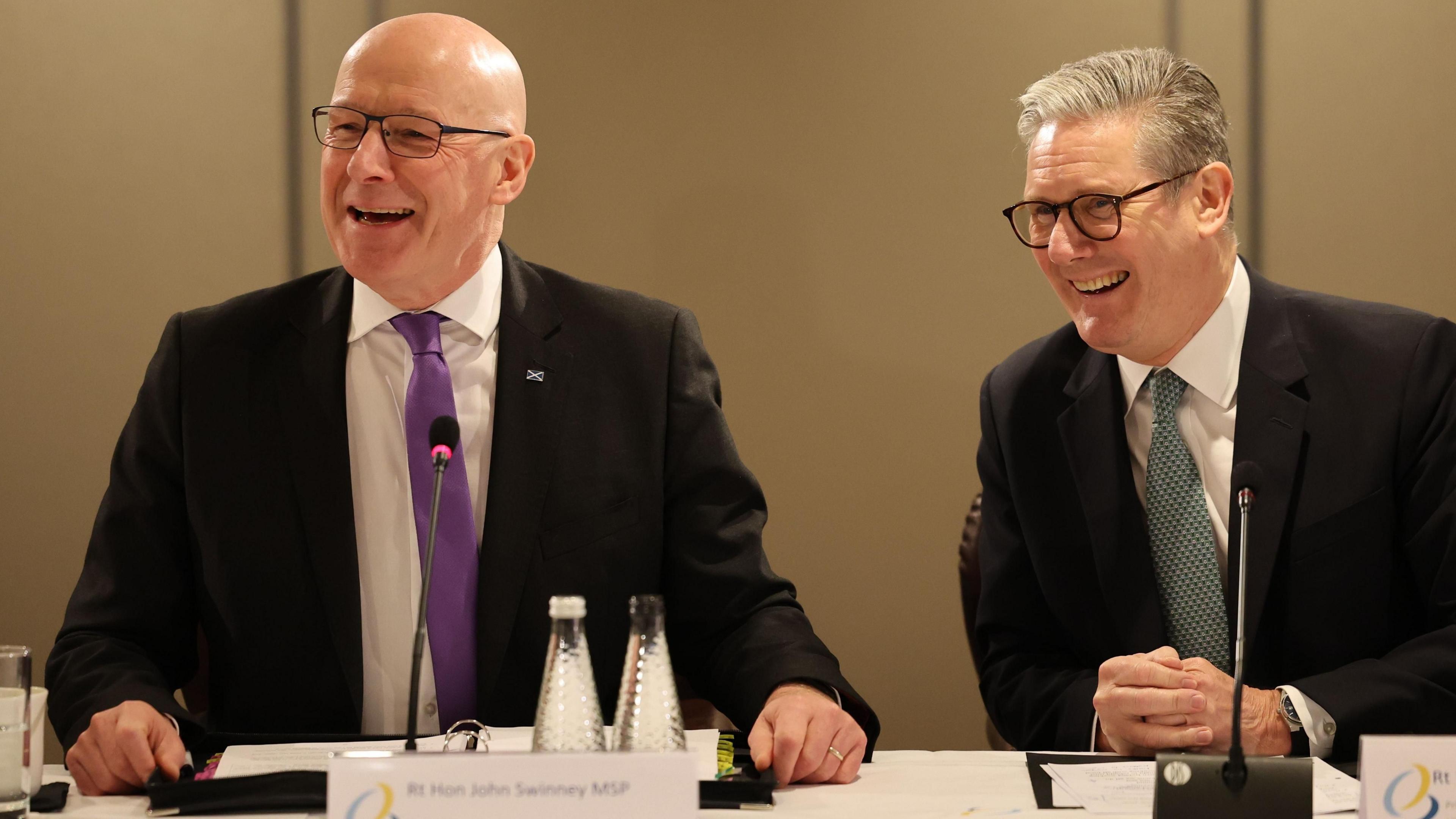 John Swinney and Sir Keir Starmer laughing while sitting at a table at the British-Irish Summit meeting in Edinburgh. Both men are wearing glasses, black shirts, white shirts and ties. There are microphones and bottles of water on the table in front of them.