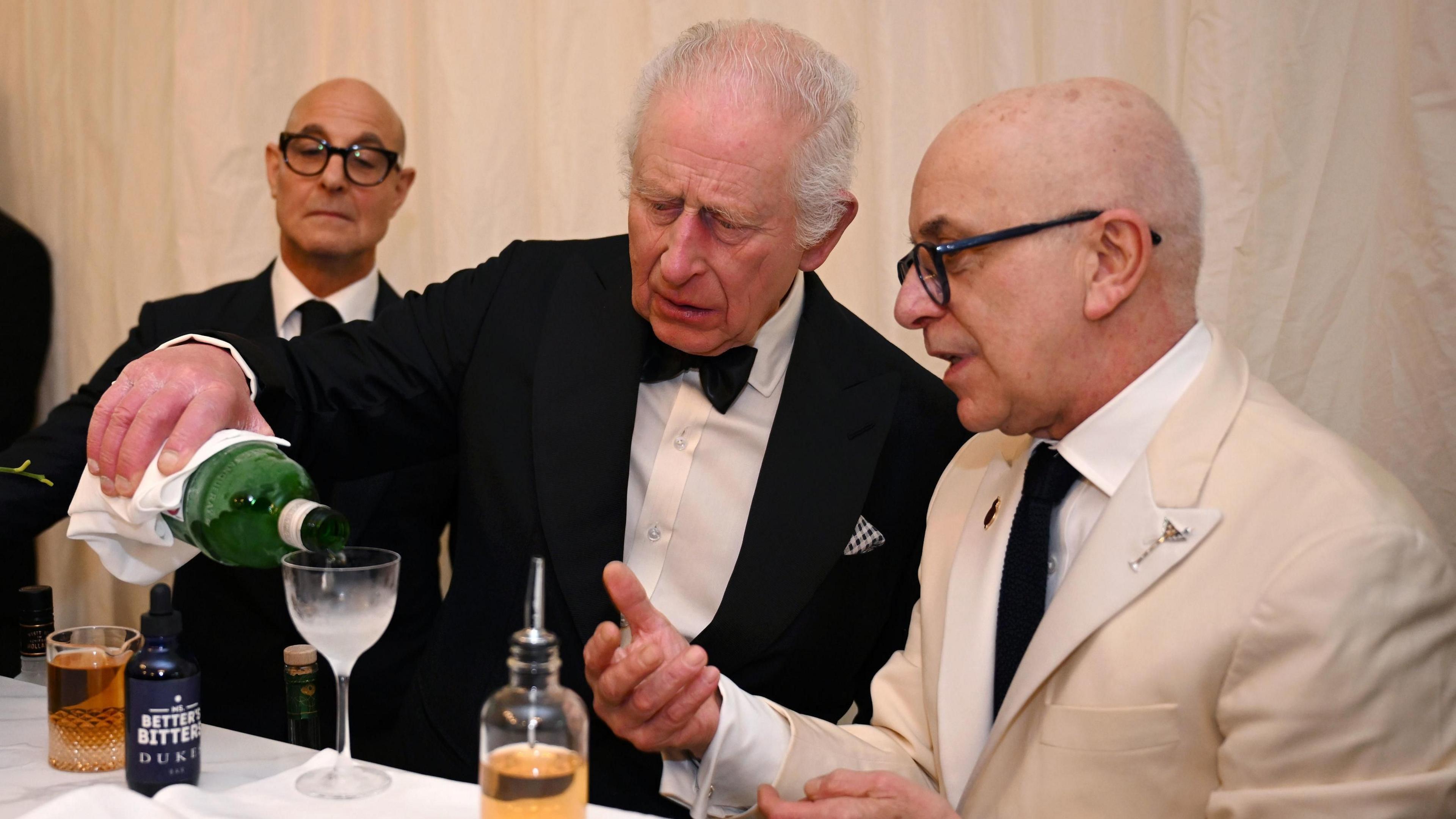 King Charles, wearing a black tuxedo, pours a drink from a green glass bottle into a small glass. Beside him sits Alessandro Palazz, who wears a cream-coloured suit with a black tie and black glasses, while Stanley Tucci watches the King over his shoulder, wearing a black suit and round black glasses.