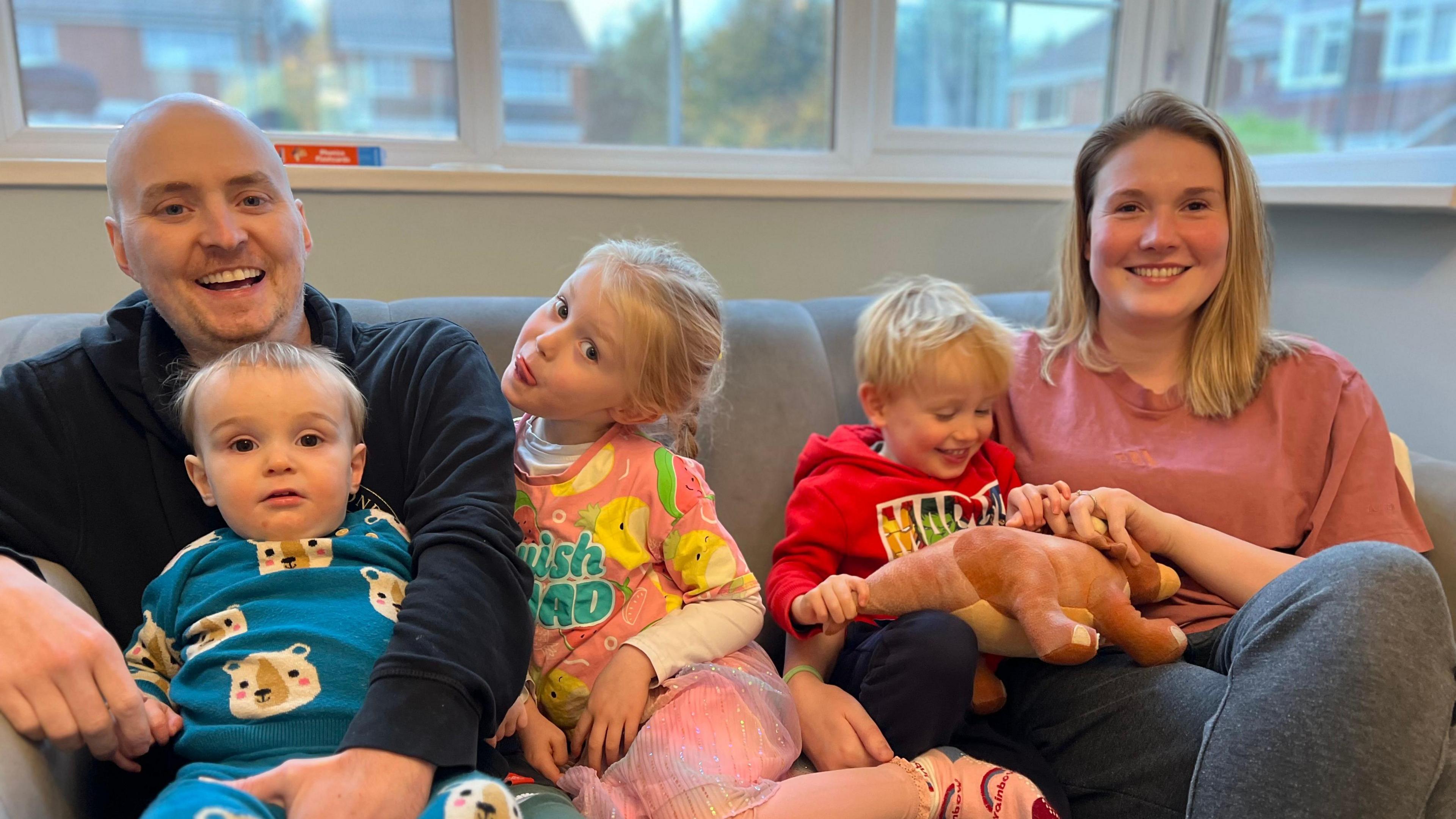 A man, far left, and a woman, far right, sat on a sofa with their three children sat in between them.