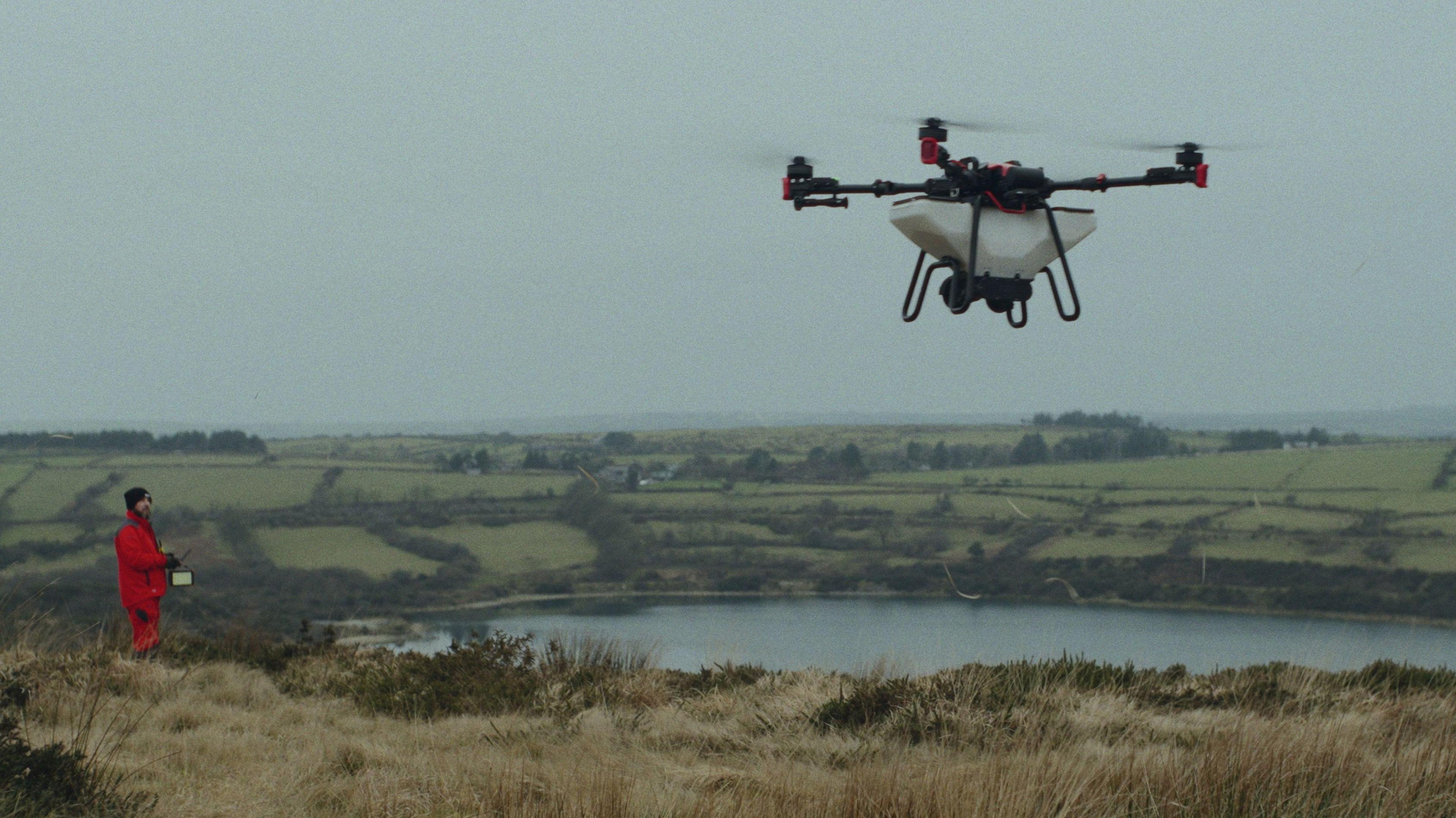 A drone with four rotors flies above the English countryside, piloted by a person wearing red overalls