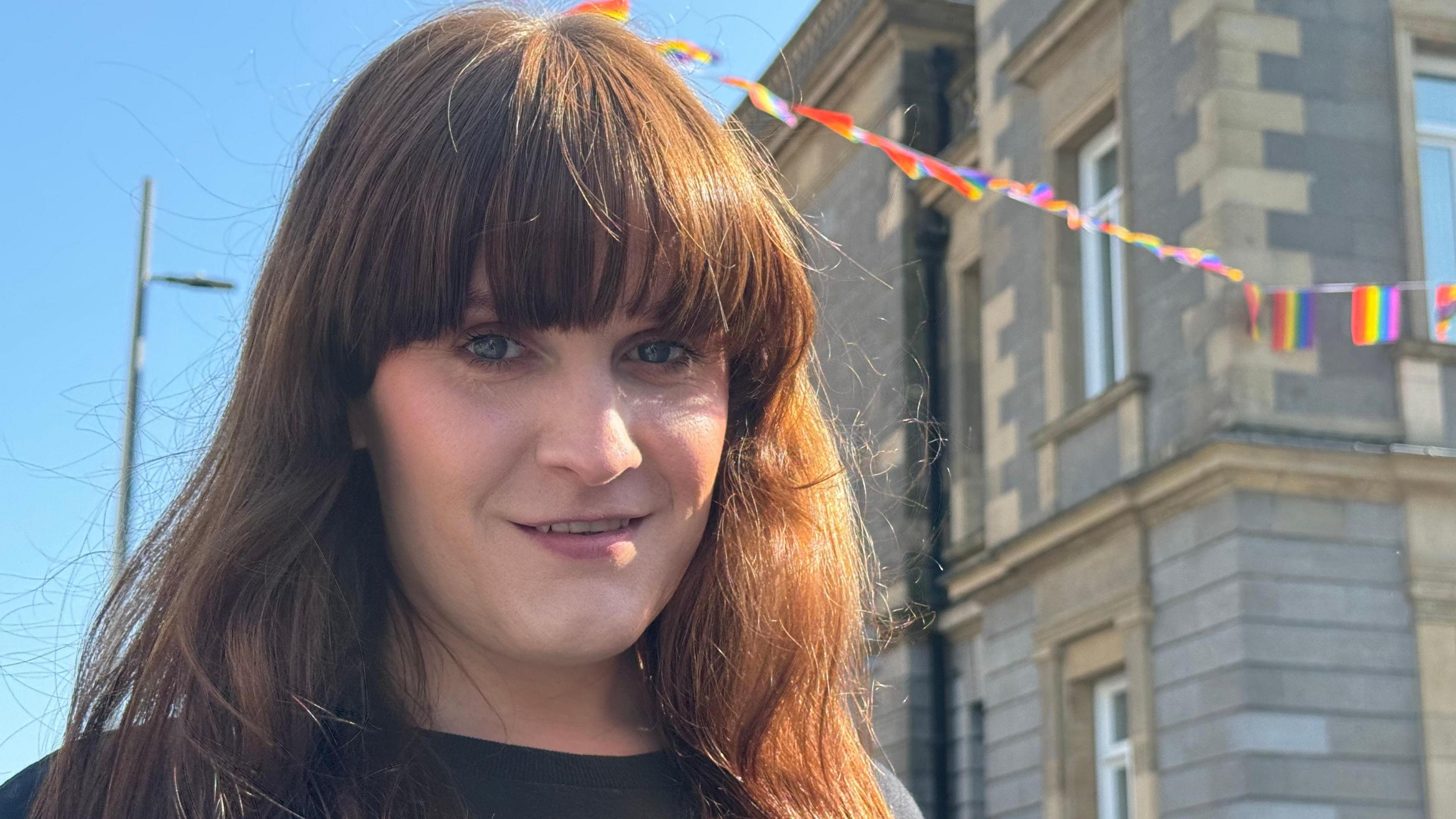 A woman with wavy brown hair and a fringe looking at the camera. Behind her hanging from a building is rainbow bunting.