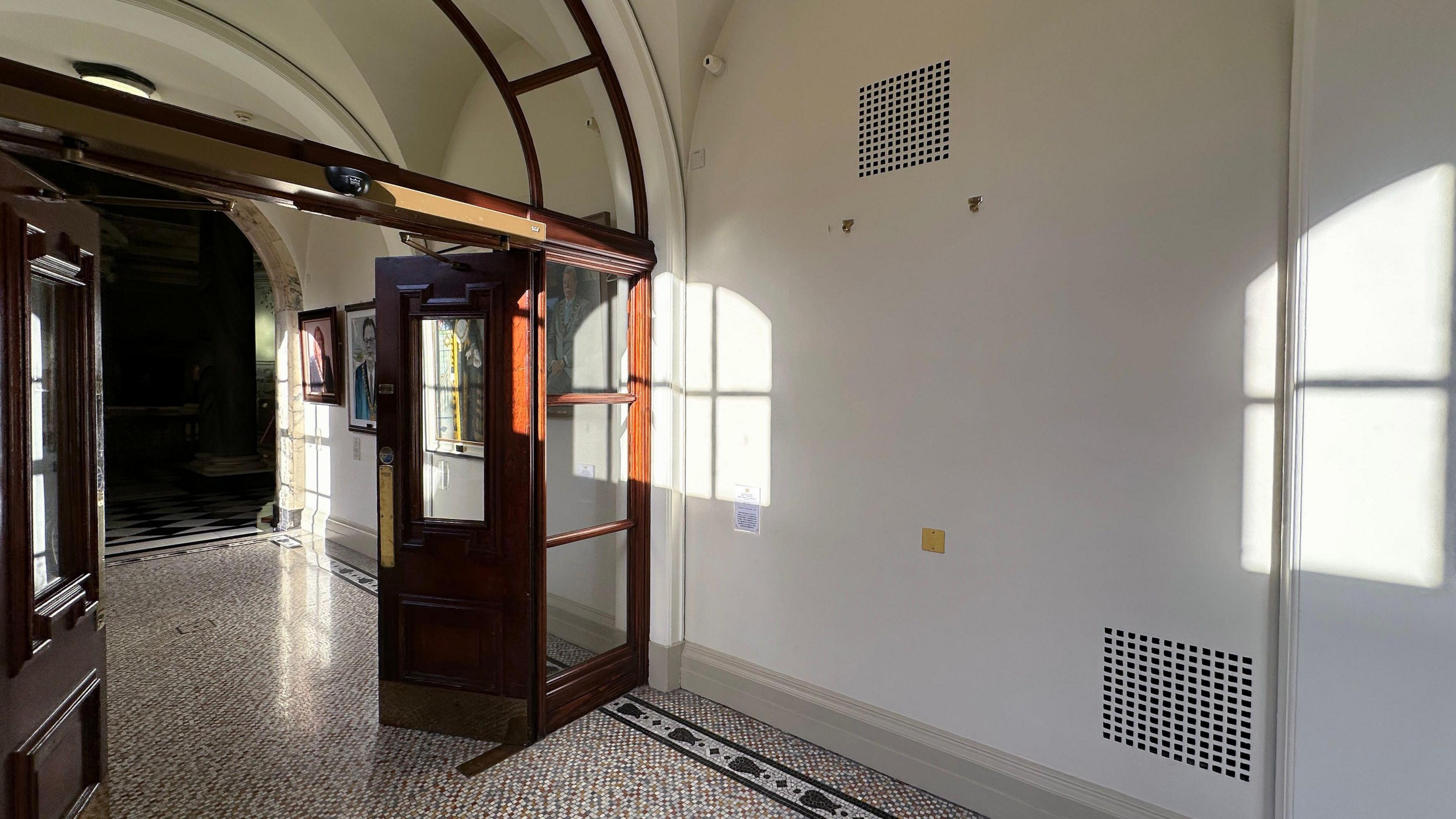 An empty space on the wall outside the banqueting hall of Belfast City Hall. There are painting hooks on the wall and a tiled floor. A brown wooden door with glass windows is open. 