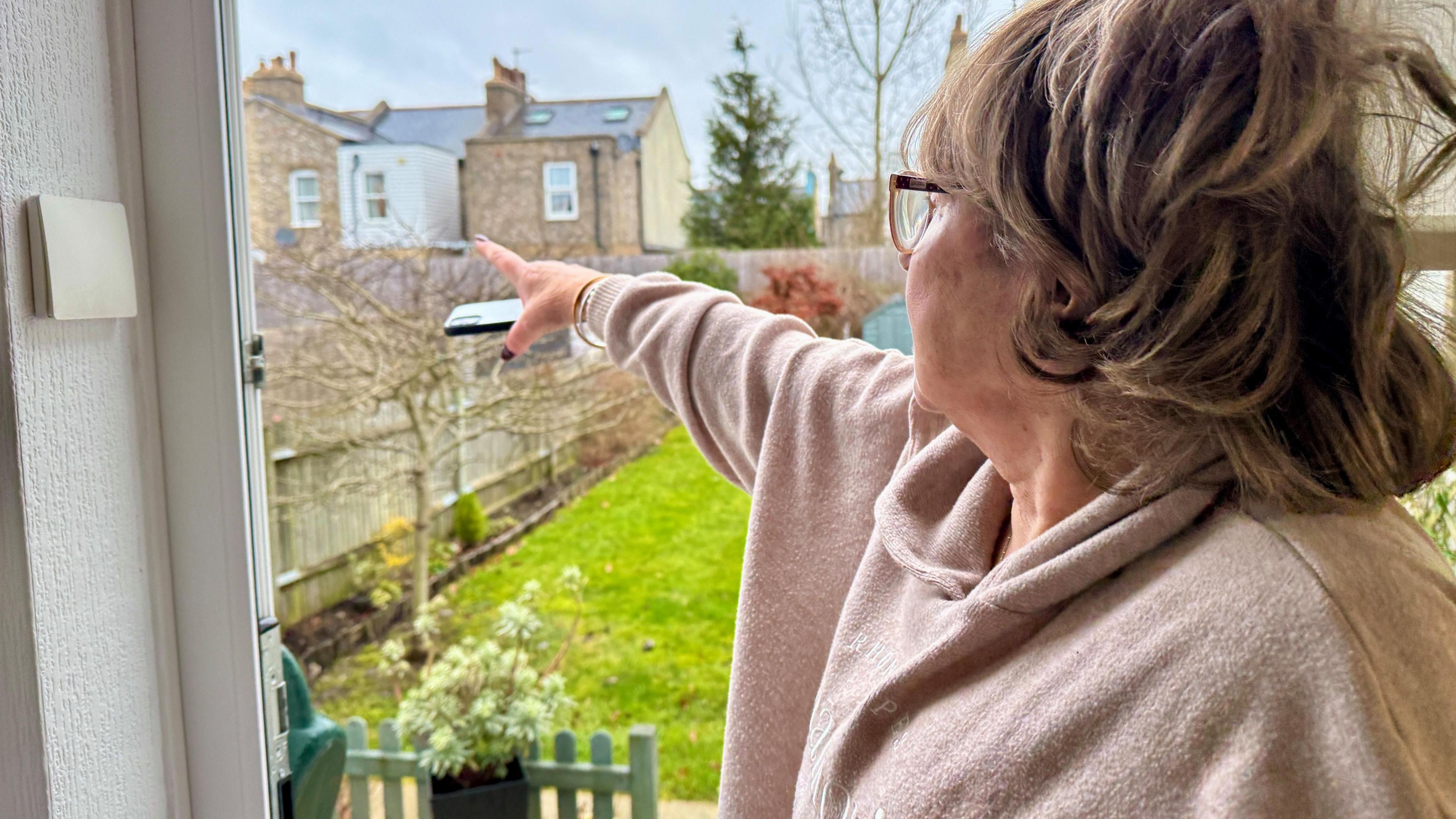 Pamela, is stood in the doorway of her back door and pointing towards the house behind her garden which are part of the EastEnders set.