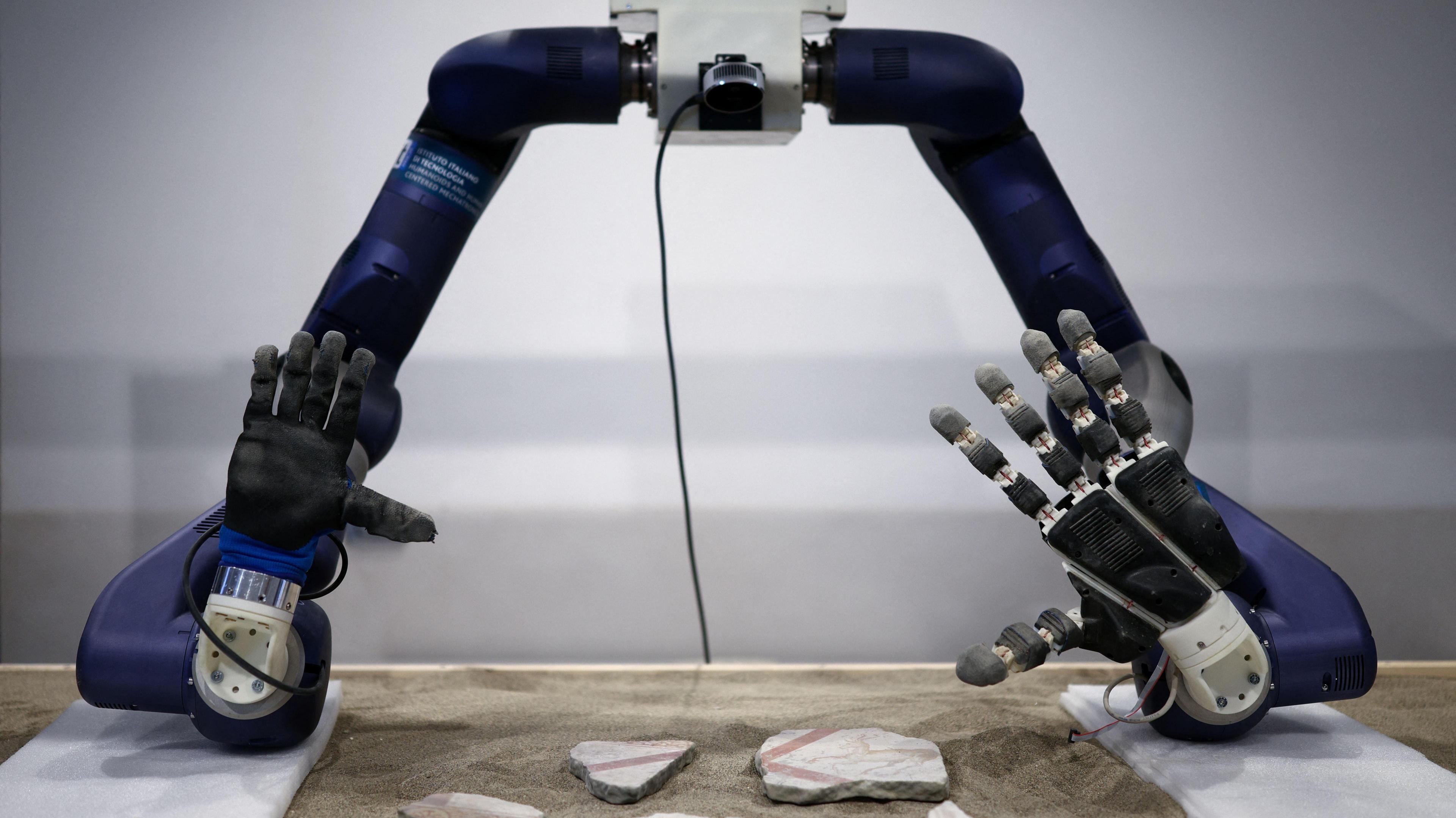 Blue arms of a robot have its palms outstretched on a table, with pieces of mosaic on the table