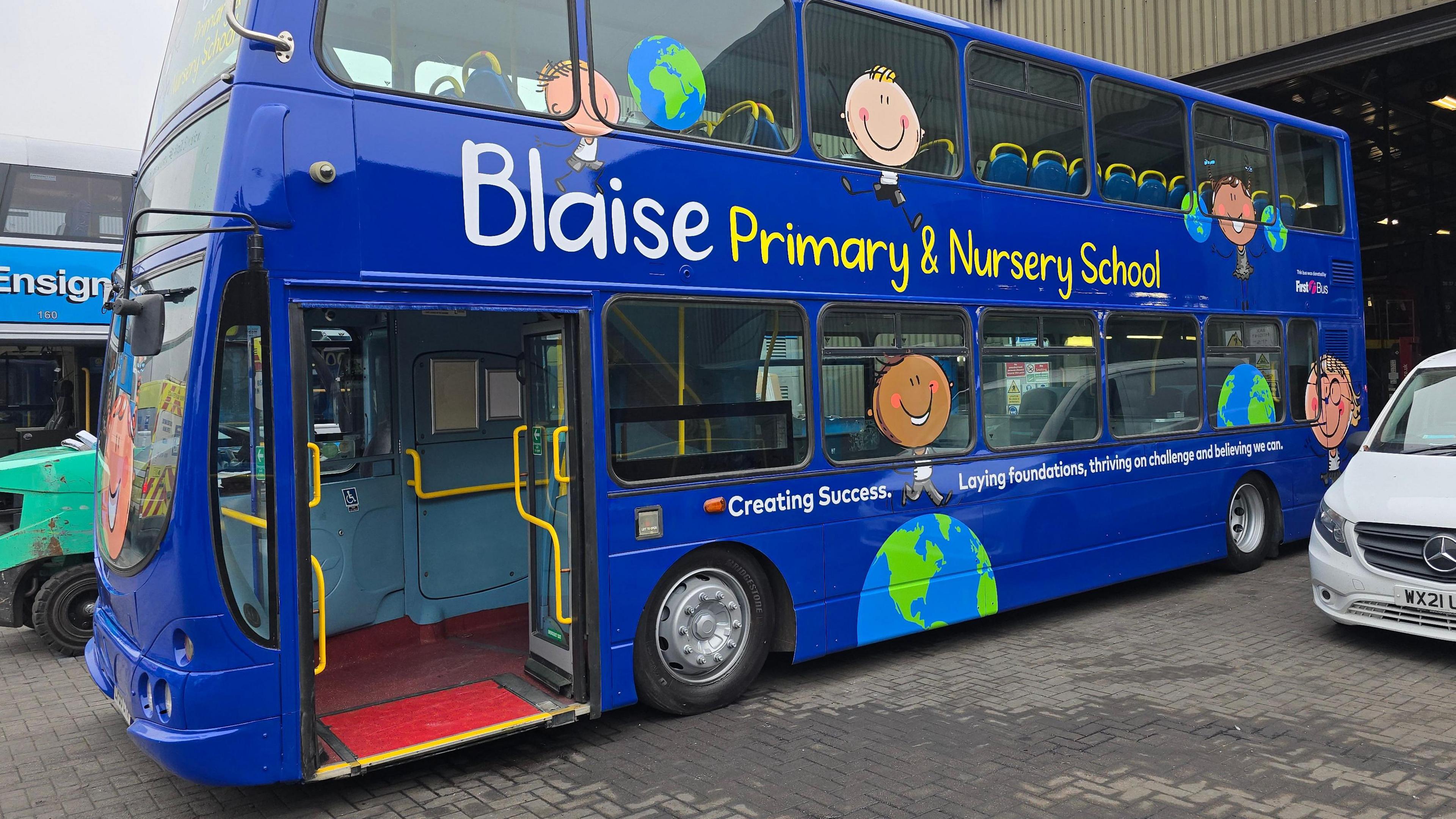 A blue, double-decker bus is parked in a depot. It has 'Blaise Primary & Nursery School' written on the side alongside drawings of children and the world. 