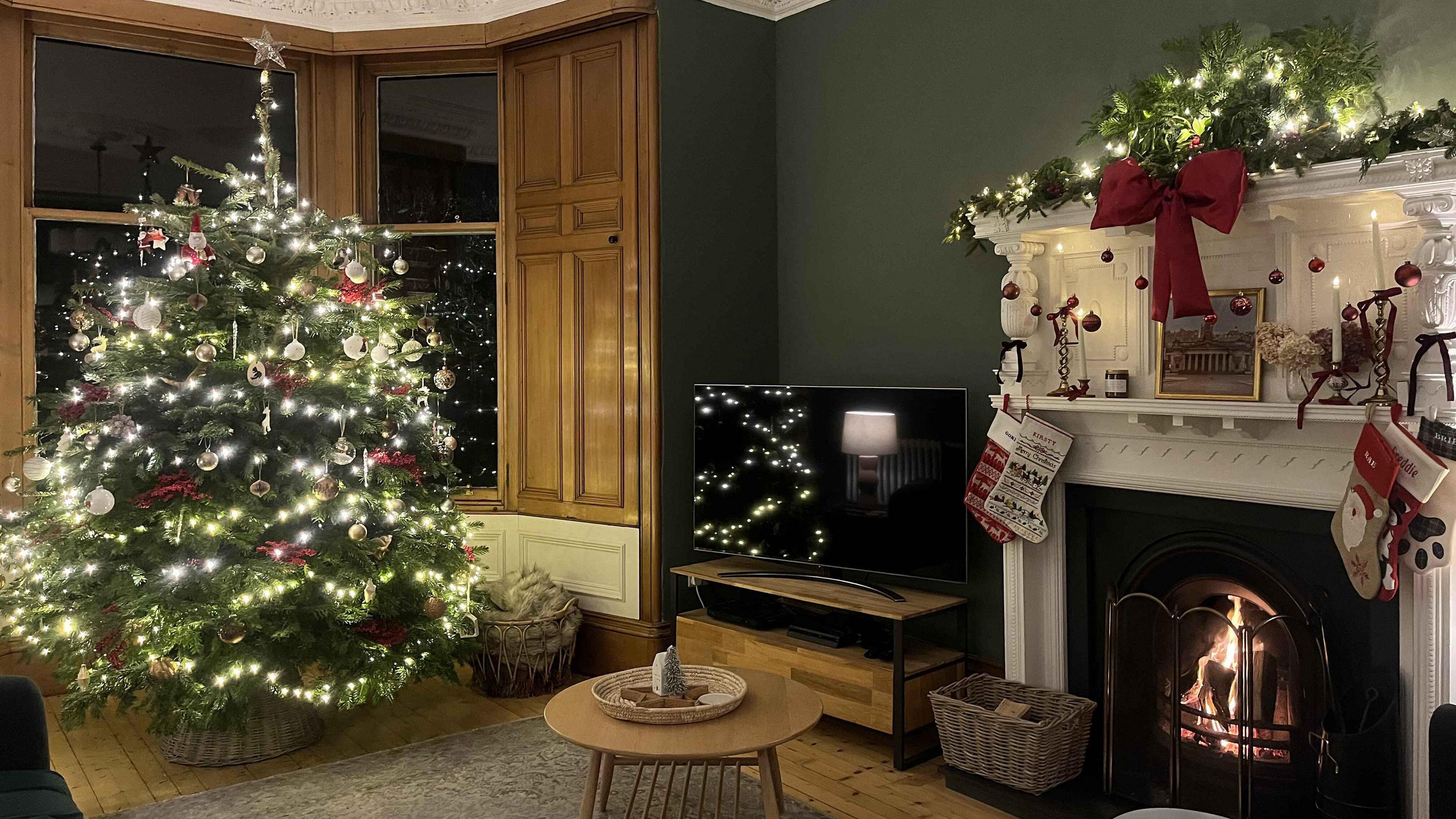 A large Christmas tree sits in front of a bay window. The tree is decorated with Christmas baubles, warm white fairy lights and a small gold star sits at the top. There is a cosy fire burning in a white fireplace, which is festooned with stockings, a garland of Christmas foliage and a large red bow. 