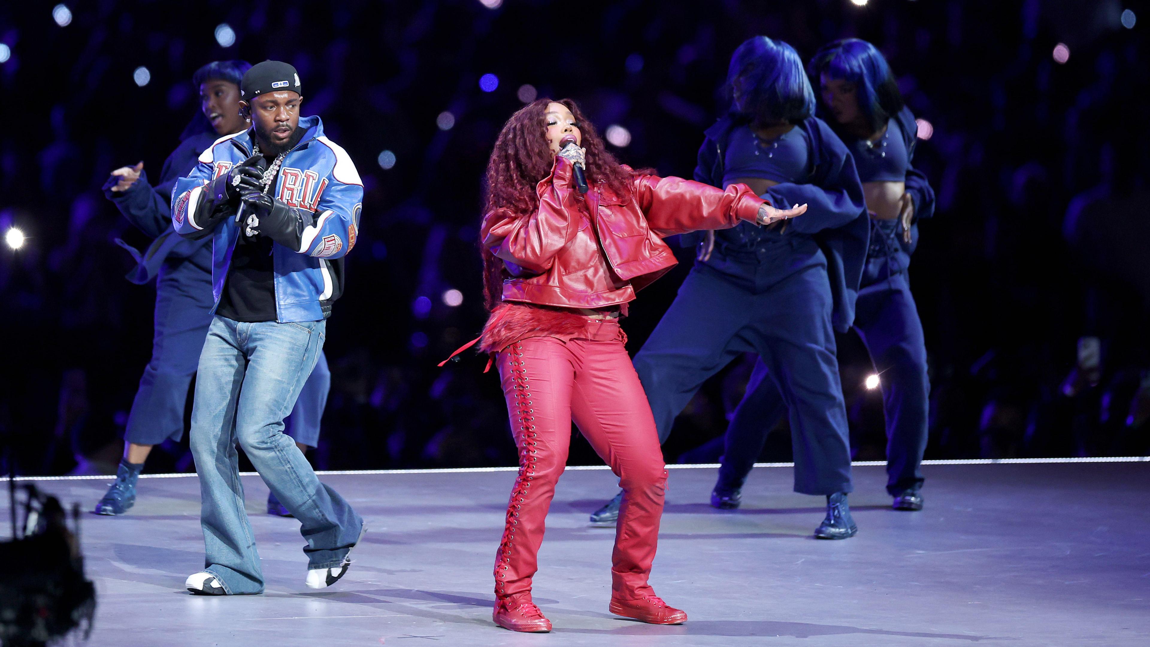 Kendrick Lamar and singer SZA perform at the Super Bowl. Kendrick Lamar is wearing a black cap, and t-shirt, a blue and black leather jacket and blue jeans. SZA is wearing red leather trousers and jacket and has long red curly hair. 