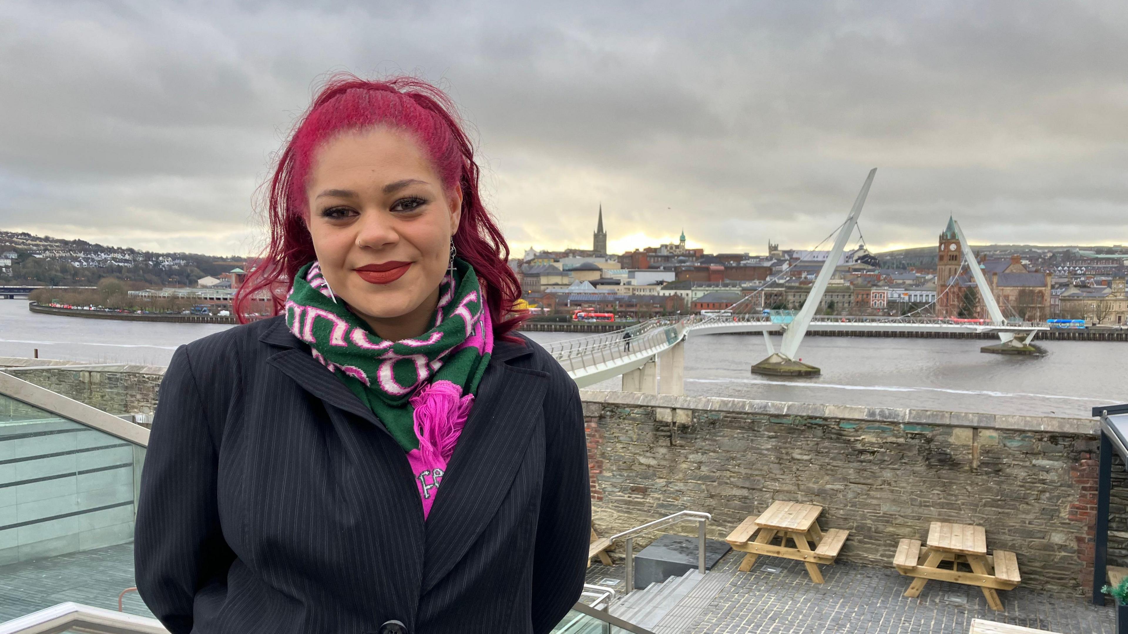 Hannah Richardson, a woman in a black suit with pink hair and a green and pink scarf. Behind her is the River Foyle and the city of Derry with the white Peace Bridge in the background. She stands immediately in front of a wall and light brown wooden picnic benches. 