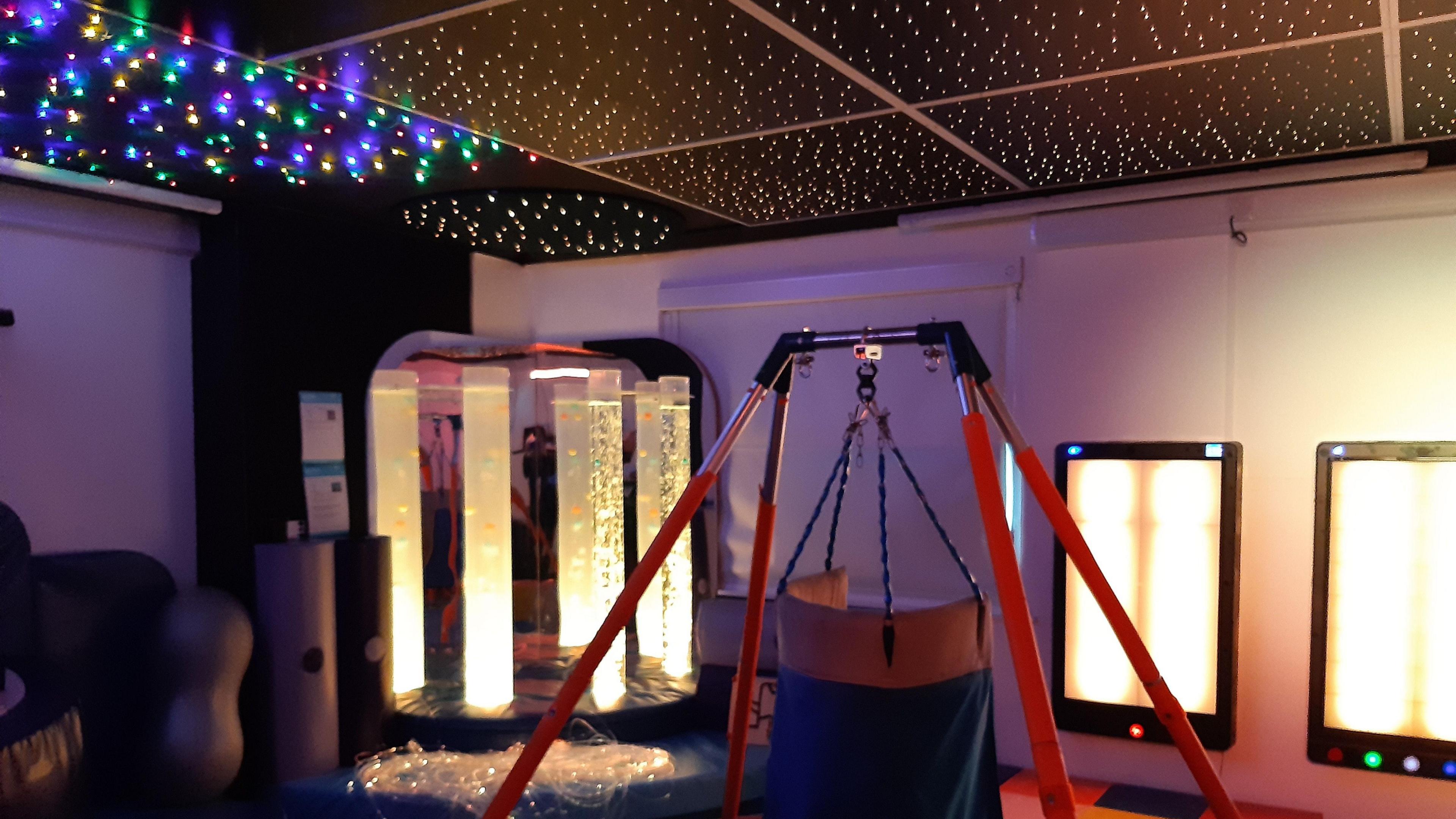 A sensory room full of lights and mirrors. The ceiling is black and filled with tiny lights like stars, some of which are multi-coloured. There is a swing with red legs and a yellow and blue seat in the middle of the room. 