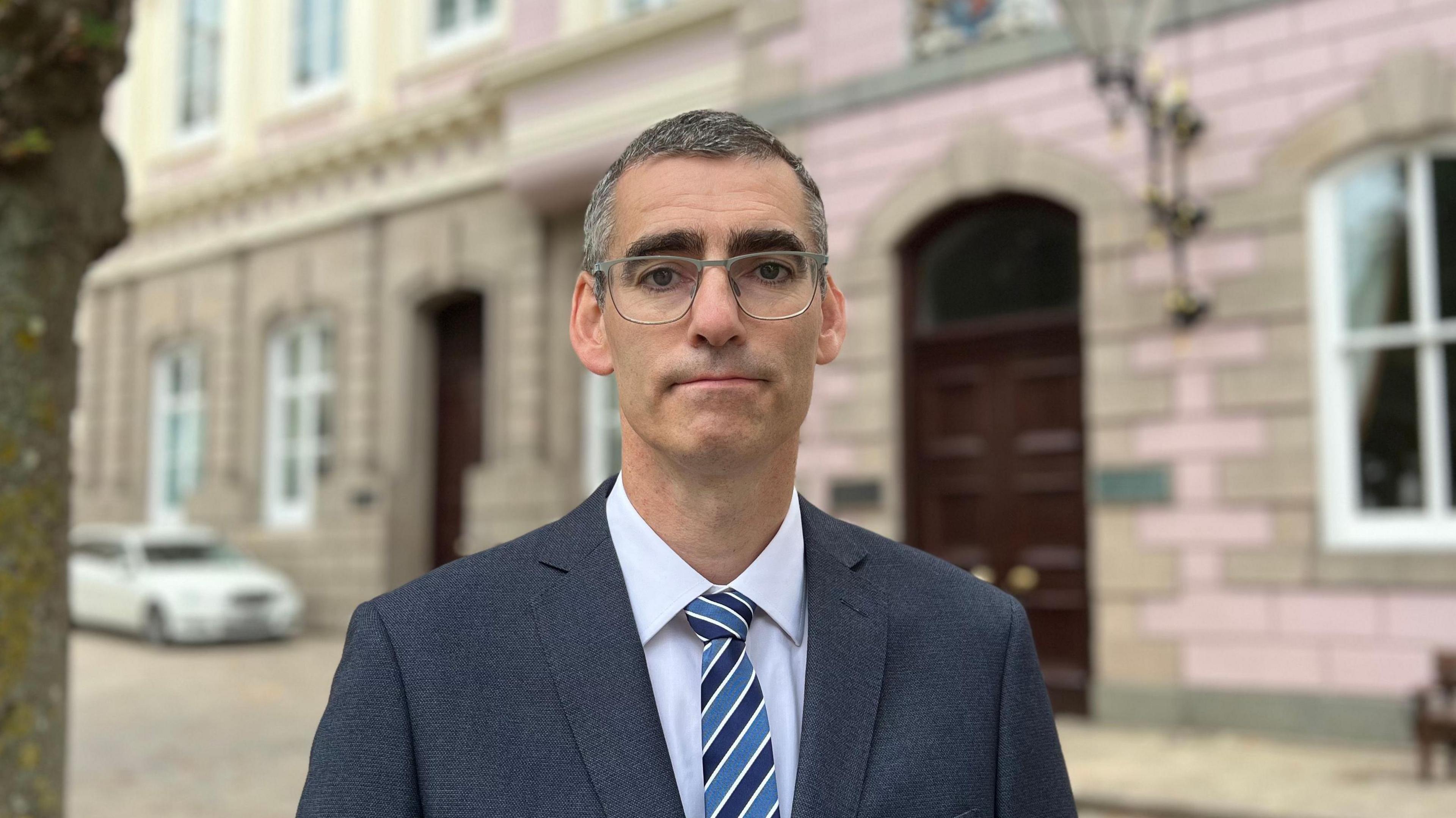 Deputy Montfort Tadier is wearing a dark blue suit, light blue shirt and blue striped tie stands in Jersey's Royal Square. A tree trunk is visible in the foreground on the left, while a bench, lantern and parked white car appear in the softly blurred background. The image is taken outdoors in daylight, with the person in sharp focus and the background out of focus.