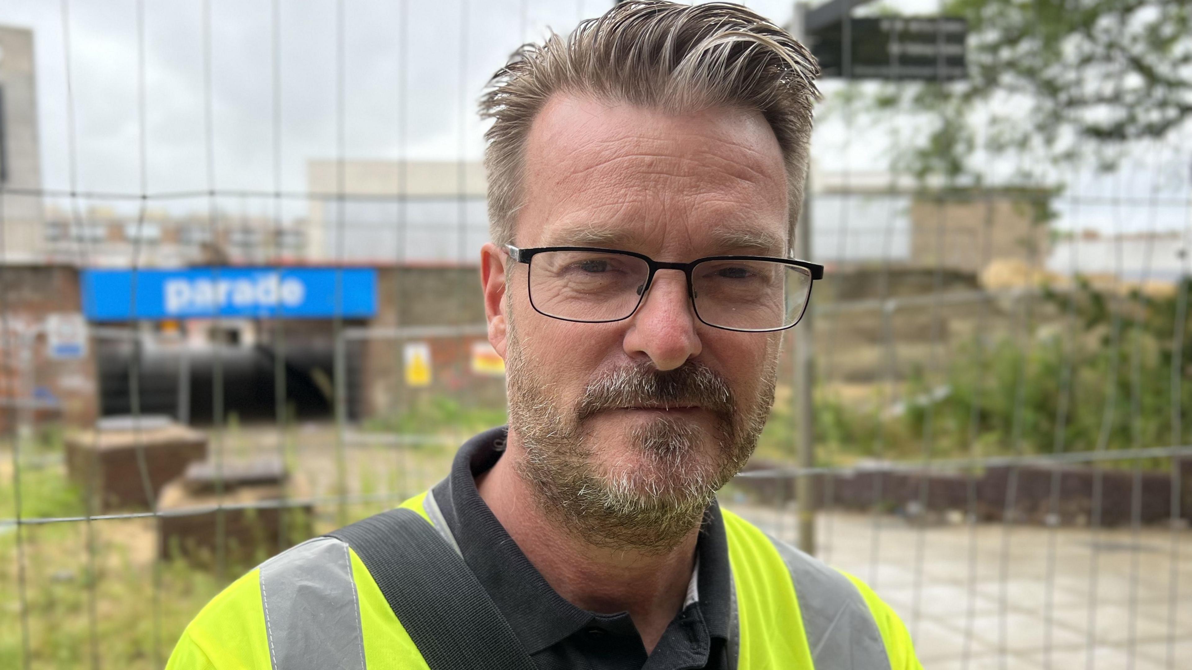 Councillor Chris Watts, a middle aged man with slicked back hair and stubble, wearing a high visibility jacket, standing in front of a fenced-off part of a town.
