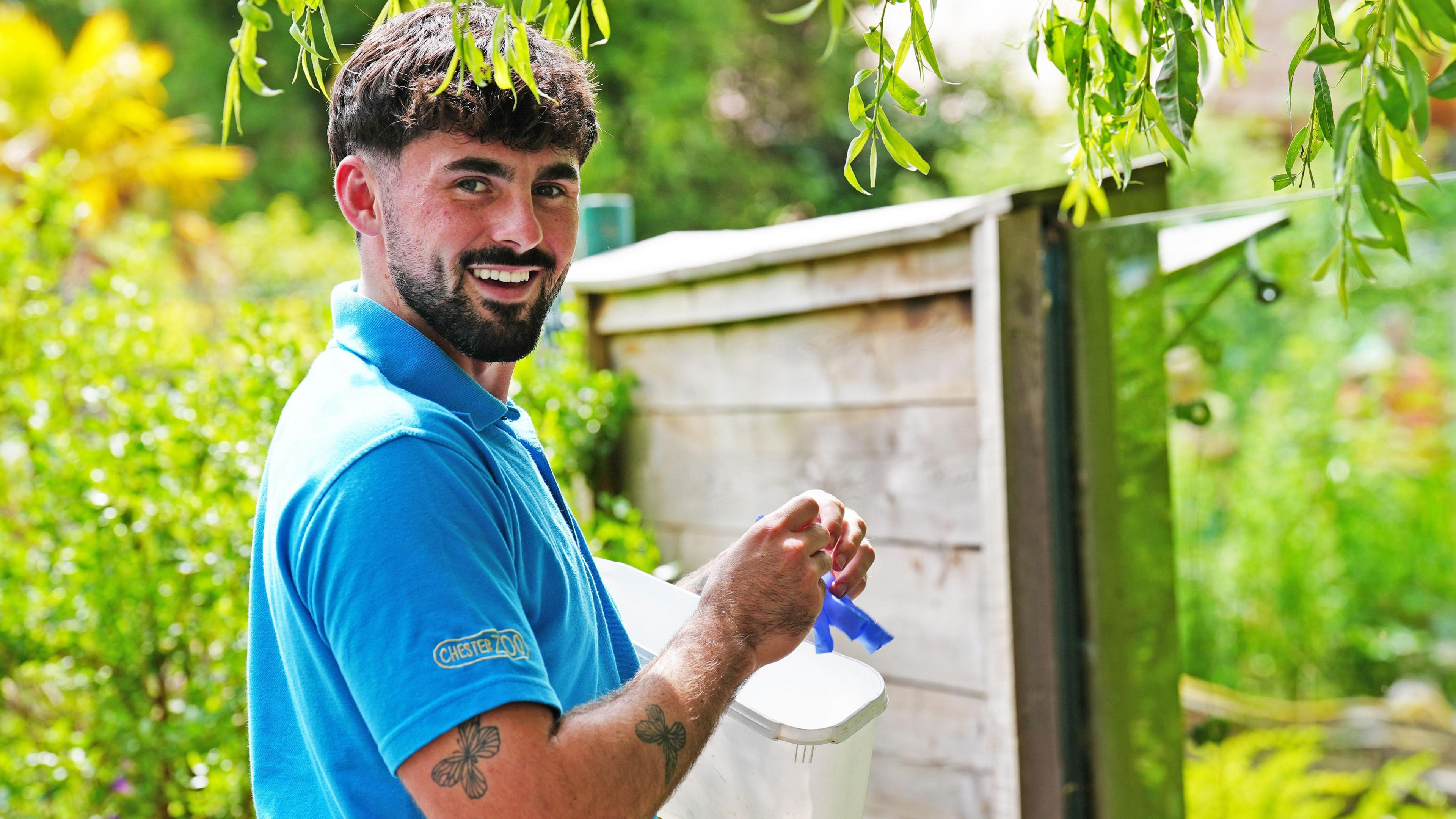 Frazer Walsh in a blue Chester Zoo branded polo shirt, holding food to feed otters. He has brown hair and is smiling at the camera, in front of a glass enclosure, surrounded by trees.