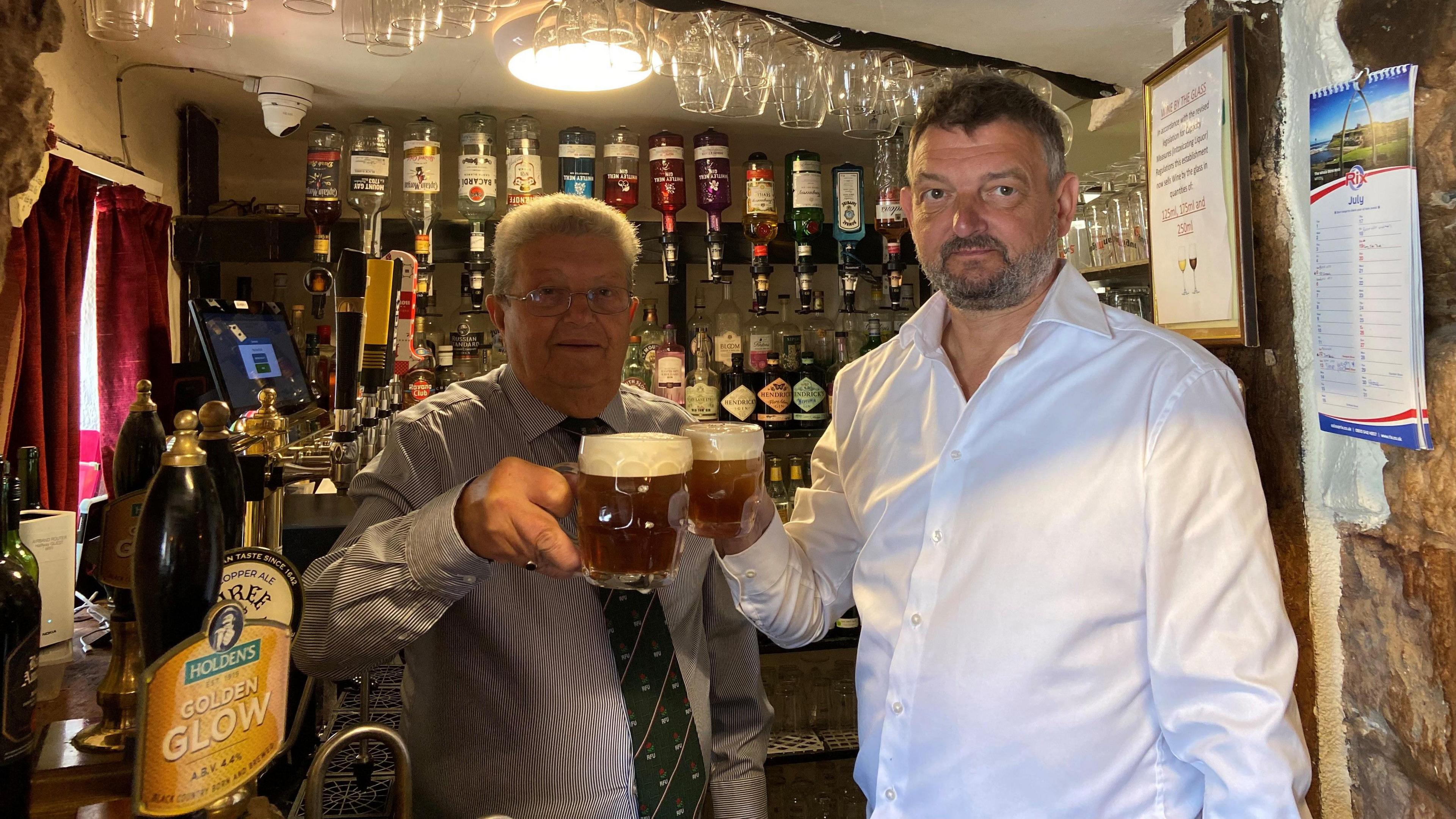 Two men are standing at a bar inside a pub. They are both holding up glass cups with beer in them. The man on the left has grey hair and glasses, and the man on the right has dark hair with grey facial hair. There are bottles in the background.