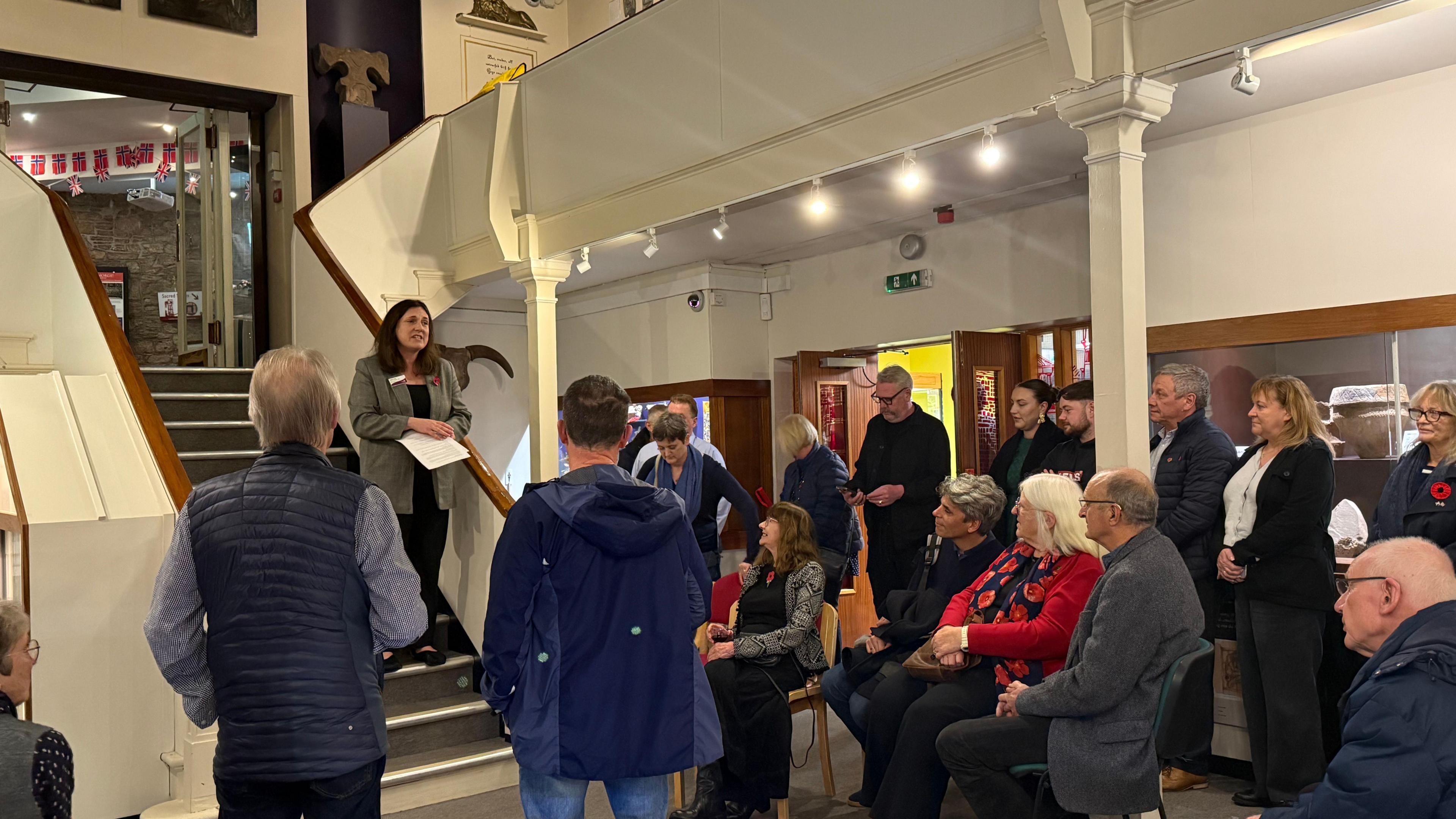 A crowd of people, some sitting and some standing, listen to a woman talk as she holds a script