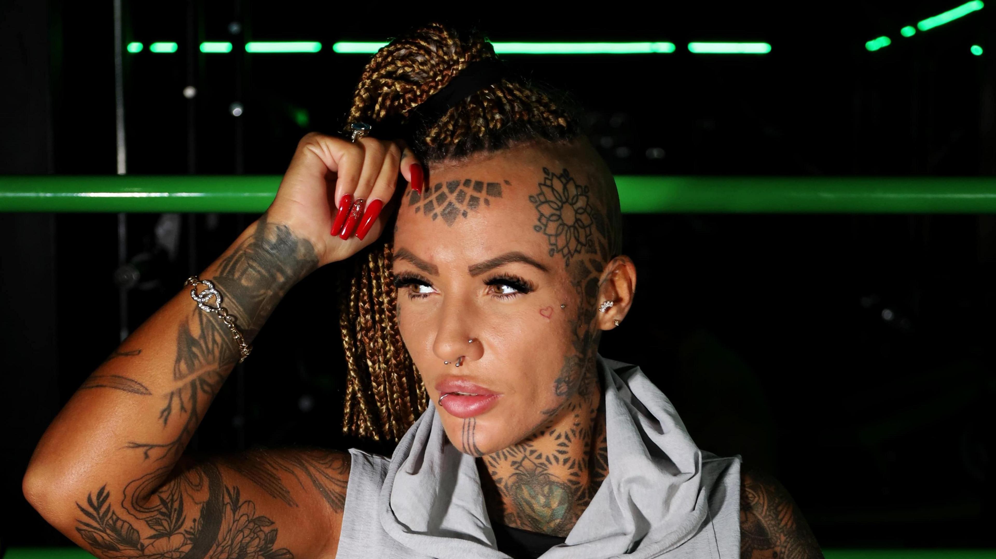 A woman with blonde braided hair and tattoos on her arms, neck and forehead pose in a sleeveless grey top.