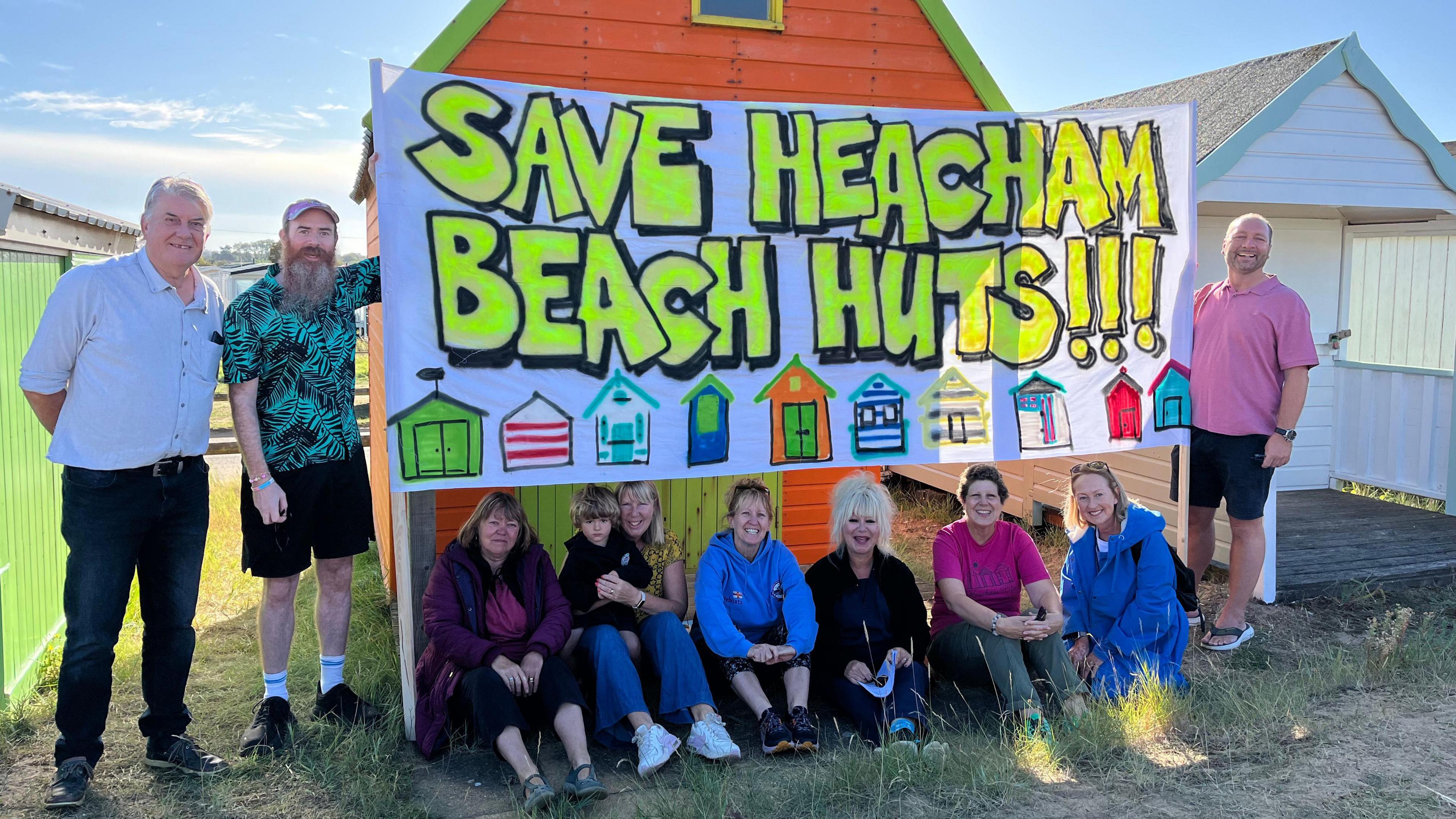 Nine people standing in front of a beach hut with a big sign above them which says "save Heacham beach huts".