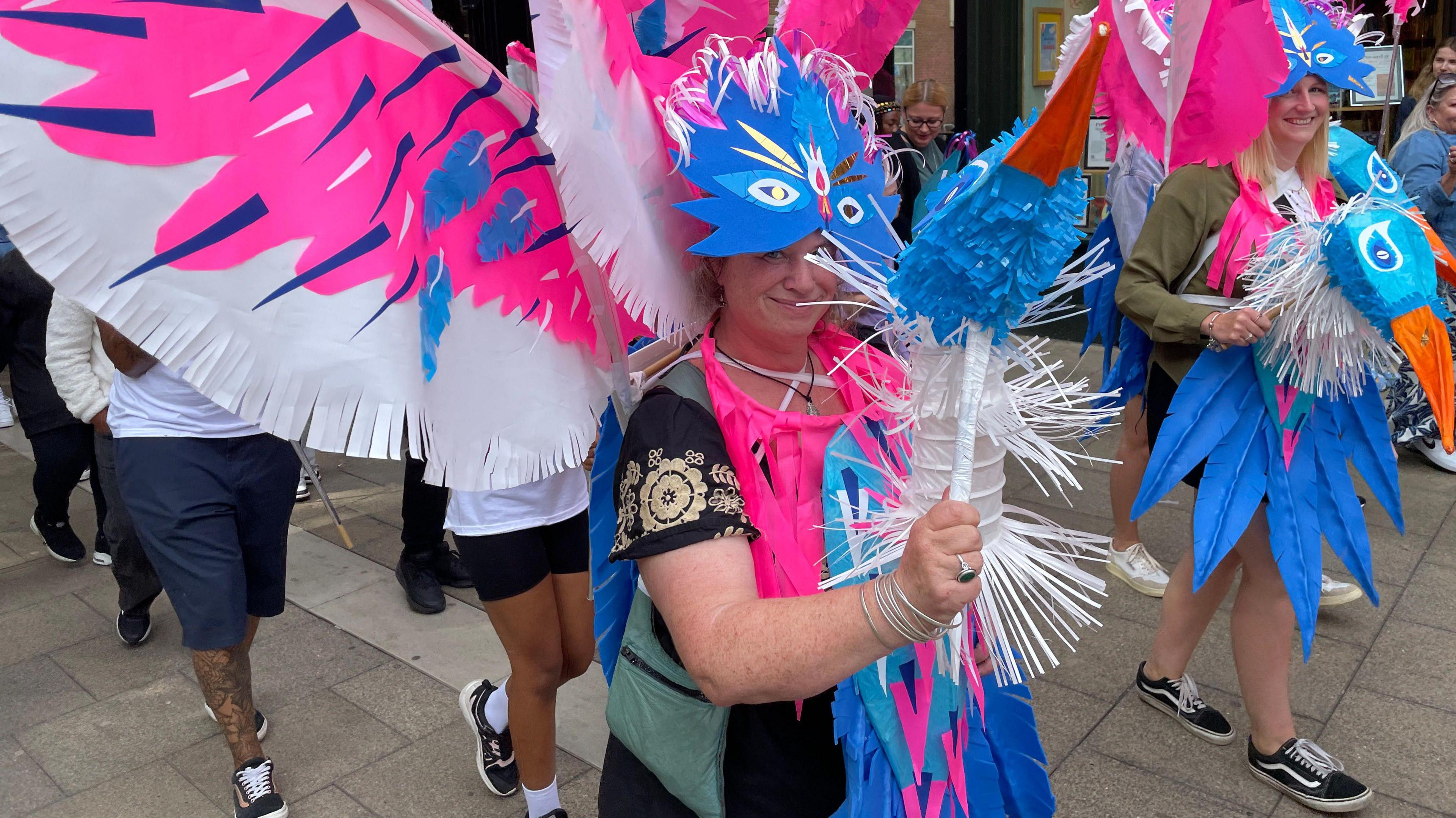 A woman in a blue, pink and white carnival costume, parading at an event, she is holding a colourful stick. People are all around her. She is smiling and looking at the camera. 