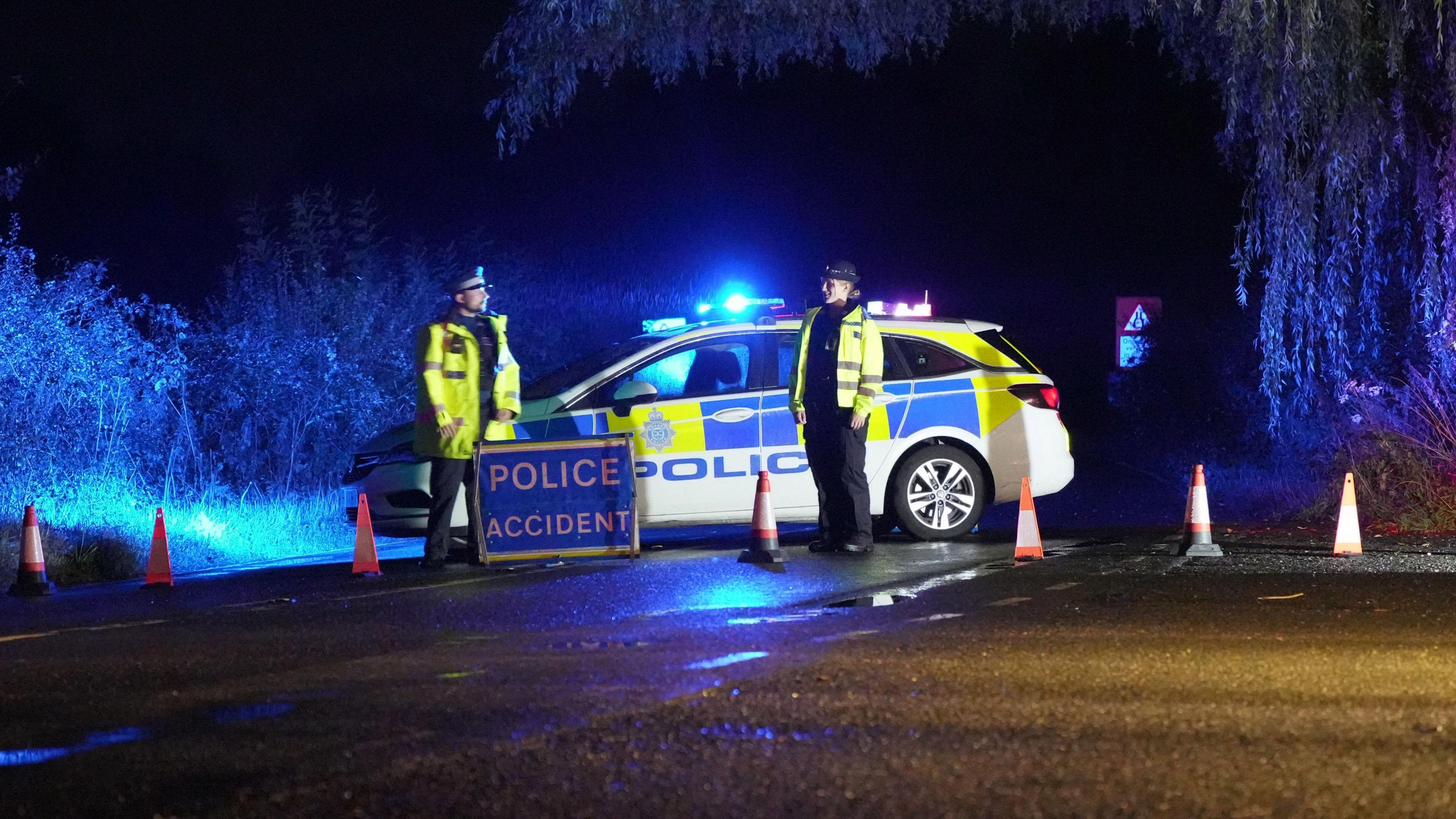 Two police officers in uniform standing in front of a police car with its lights on next to a sign that reads "police accident".