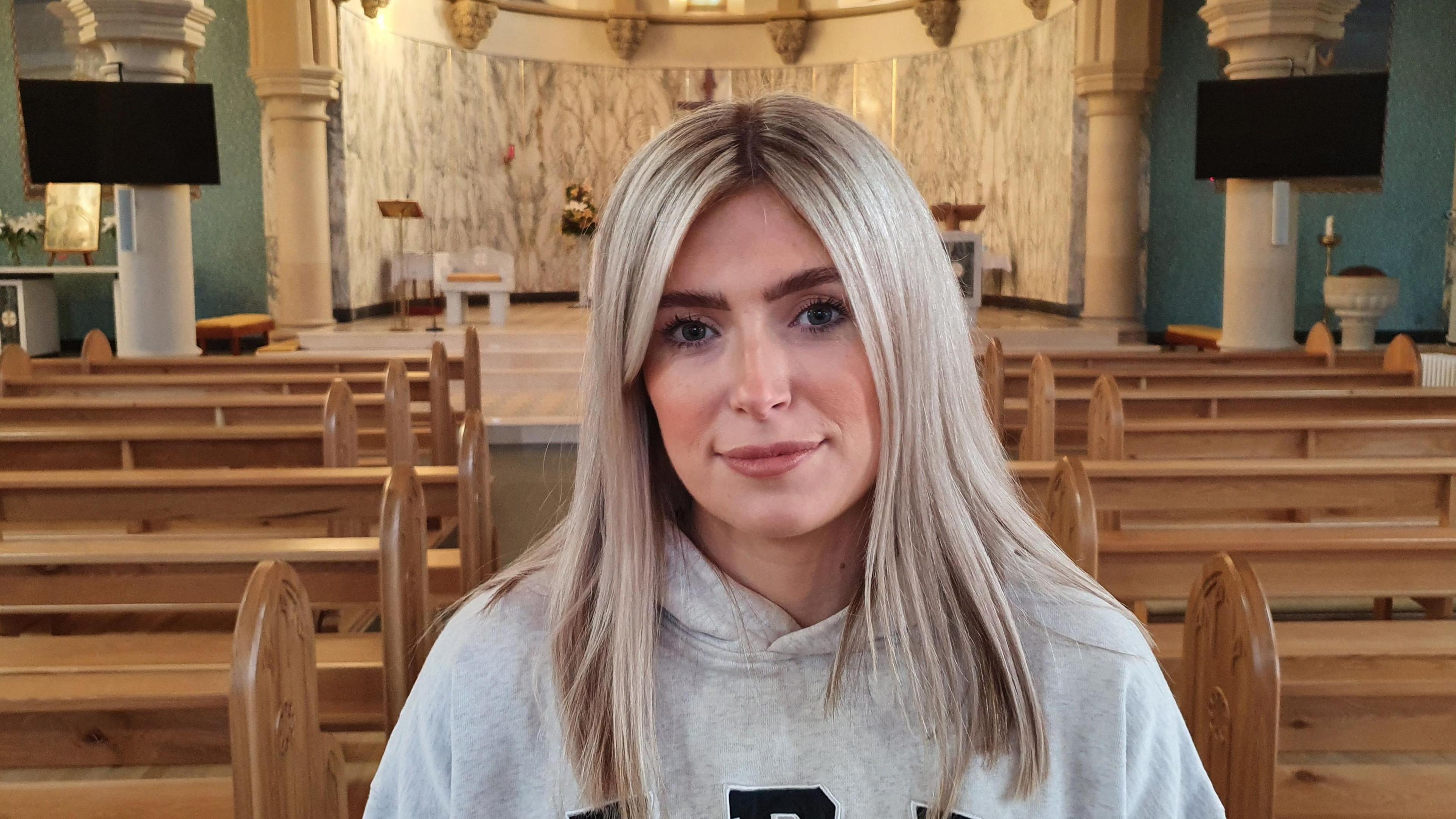 Siobhan McGowan standing in  church  with pews behind her. She has long, straight, blonde hair and is wearing makeup and a grey hooded sweatshirt.