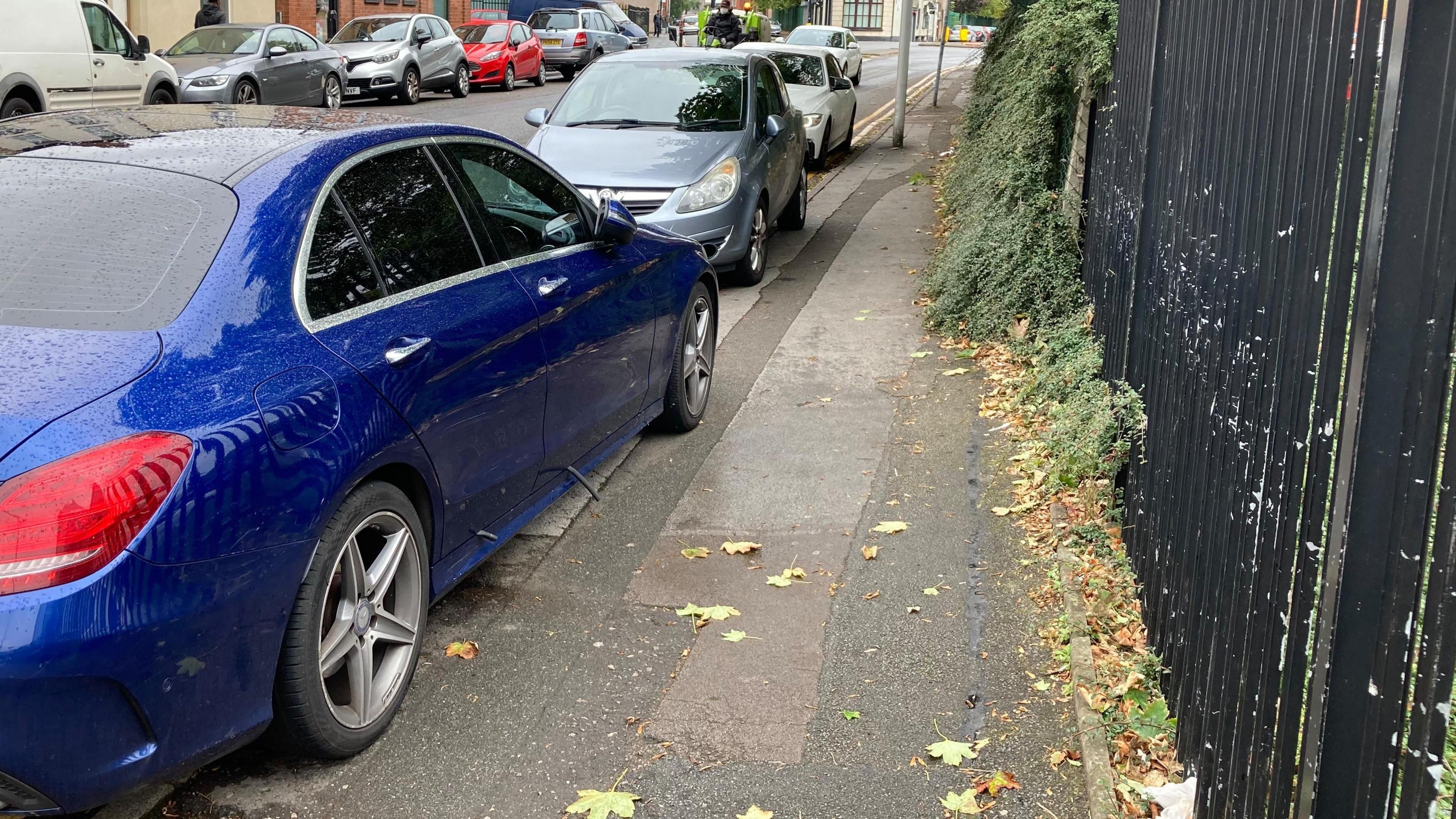 Blue and grey cars parked with their wheels on the pavement. There are metal railings on the other side of the pavement.