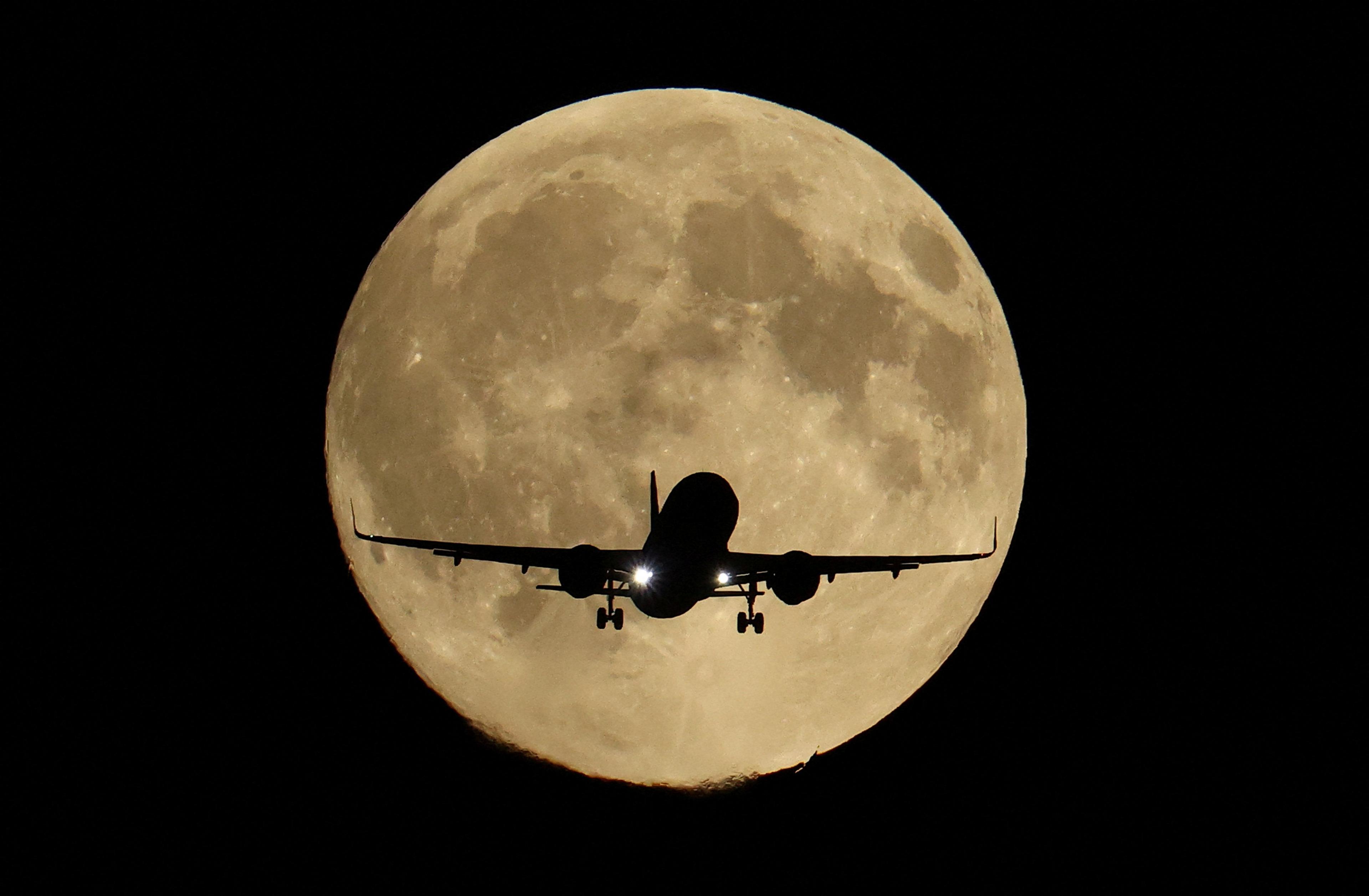 A passenger plane appears in silhouette in front of a full moon in a black night sky