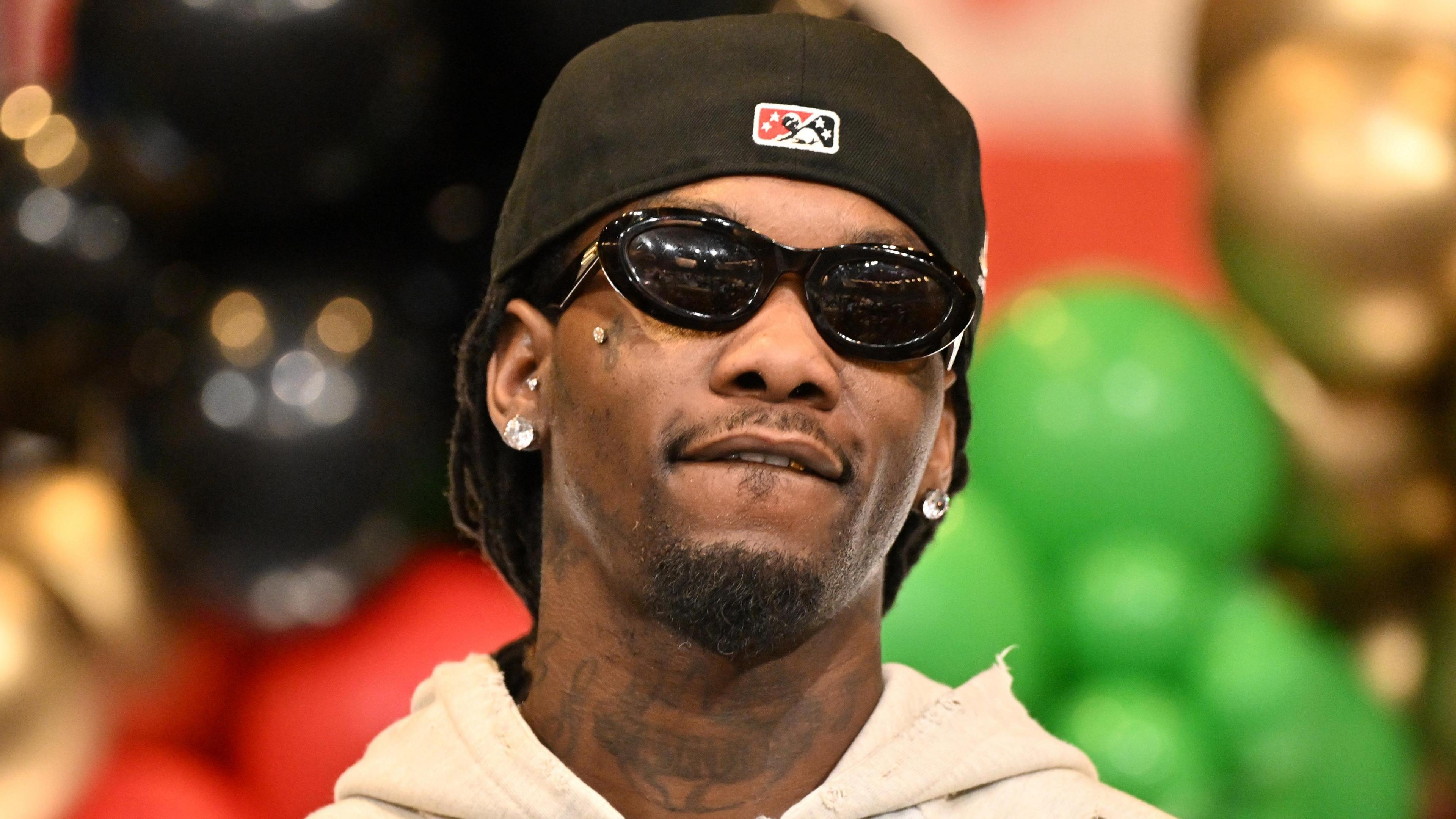 Offset in December 2024. He wears a black cap twisted back, dark sunglasses, and a zip-up beige hoody. He has a tattooed neck, a cropped beard and diamond earrings. He's pictured in front of a display, made up of black, green, gold and red balloons. 