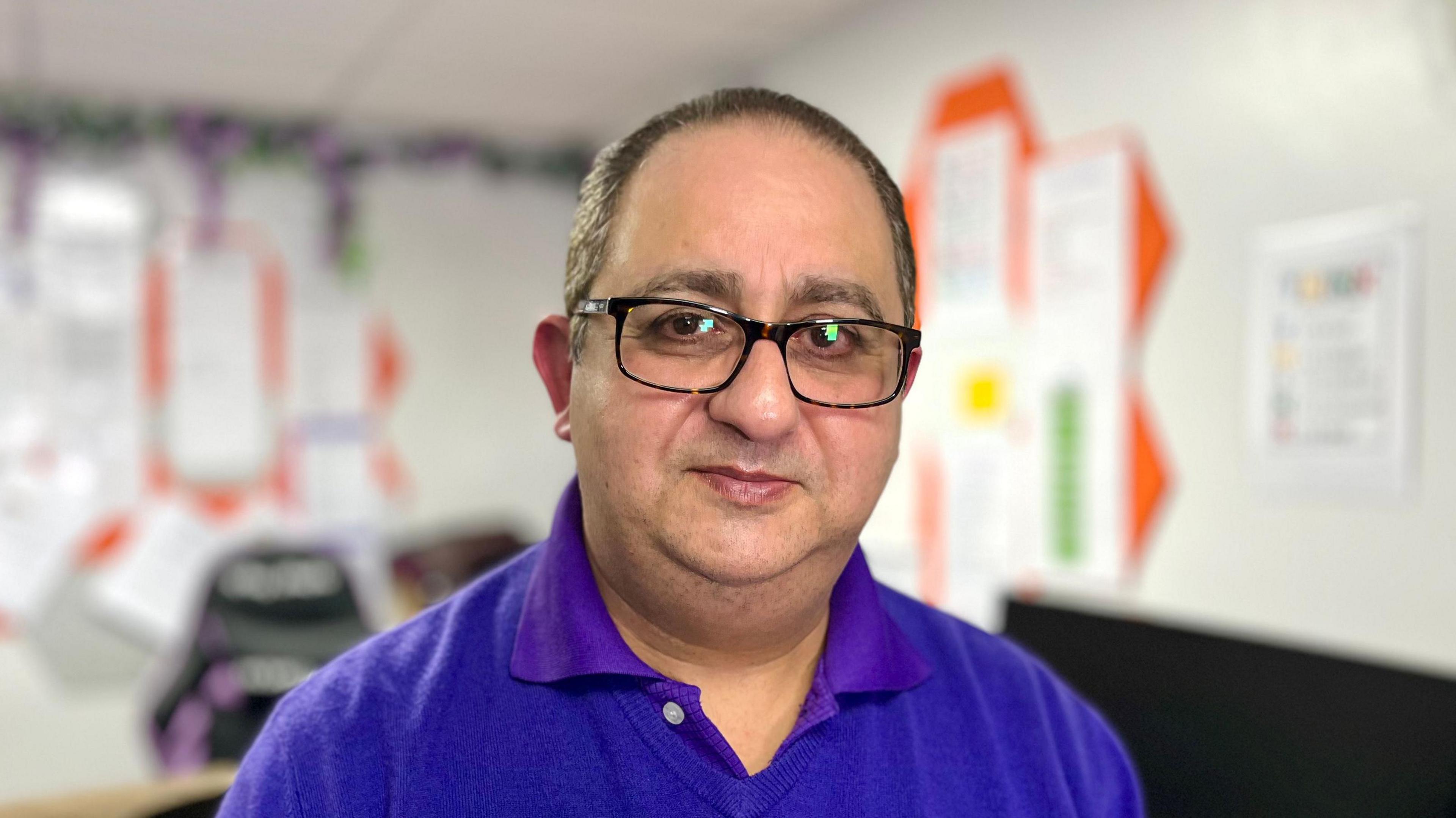 A man in wearing glasses and a purple care home shirt sits at an office desk.