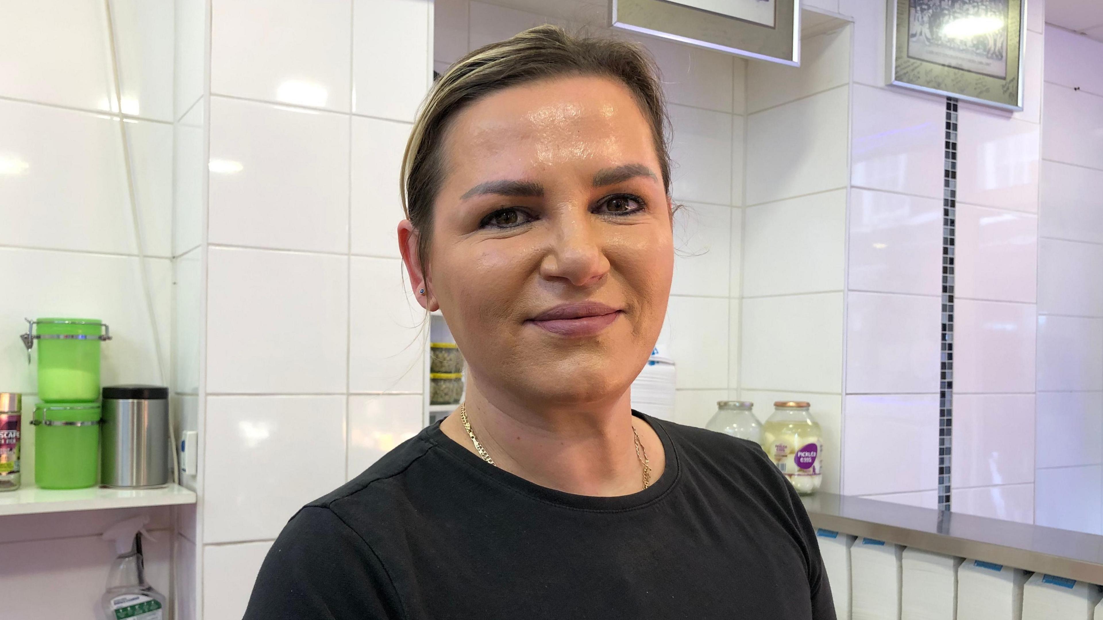 Dorina Gjoshi standing behind a fish shop counter against a white tiled wall background wearing a black top and hair tied back. 