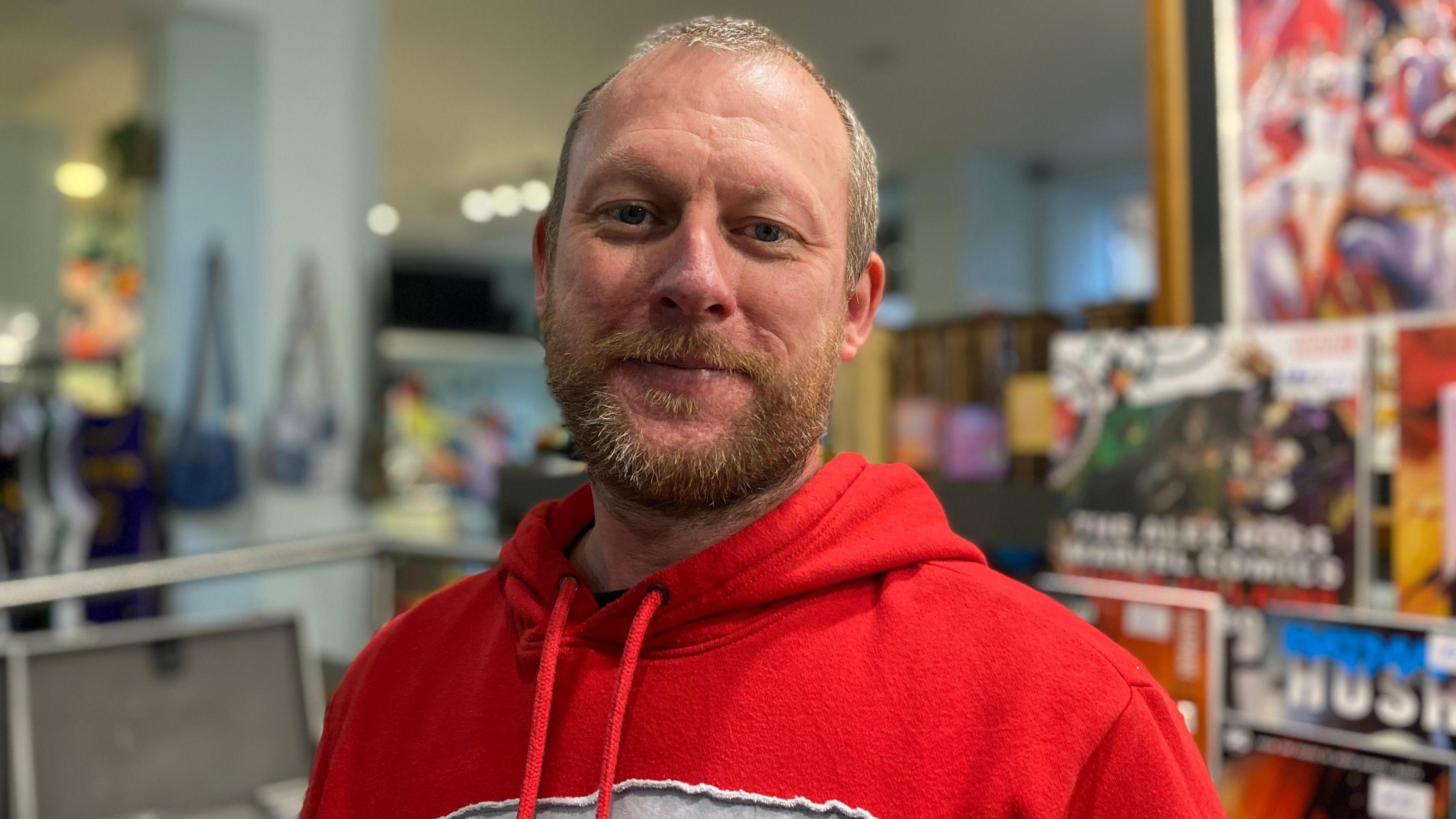 Lee Evans has short brown hair and is wearing a red hoodie. He is standing inside the vintage shop filled with clothes, bags and posters