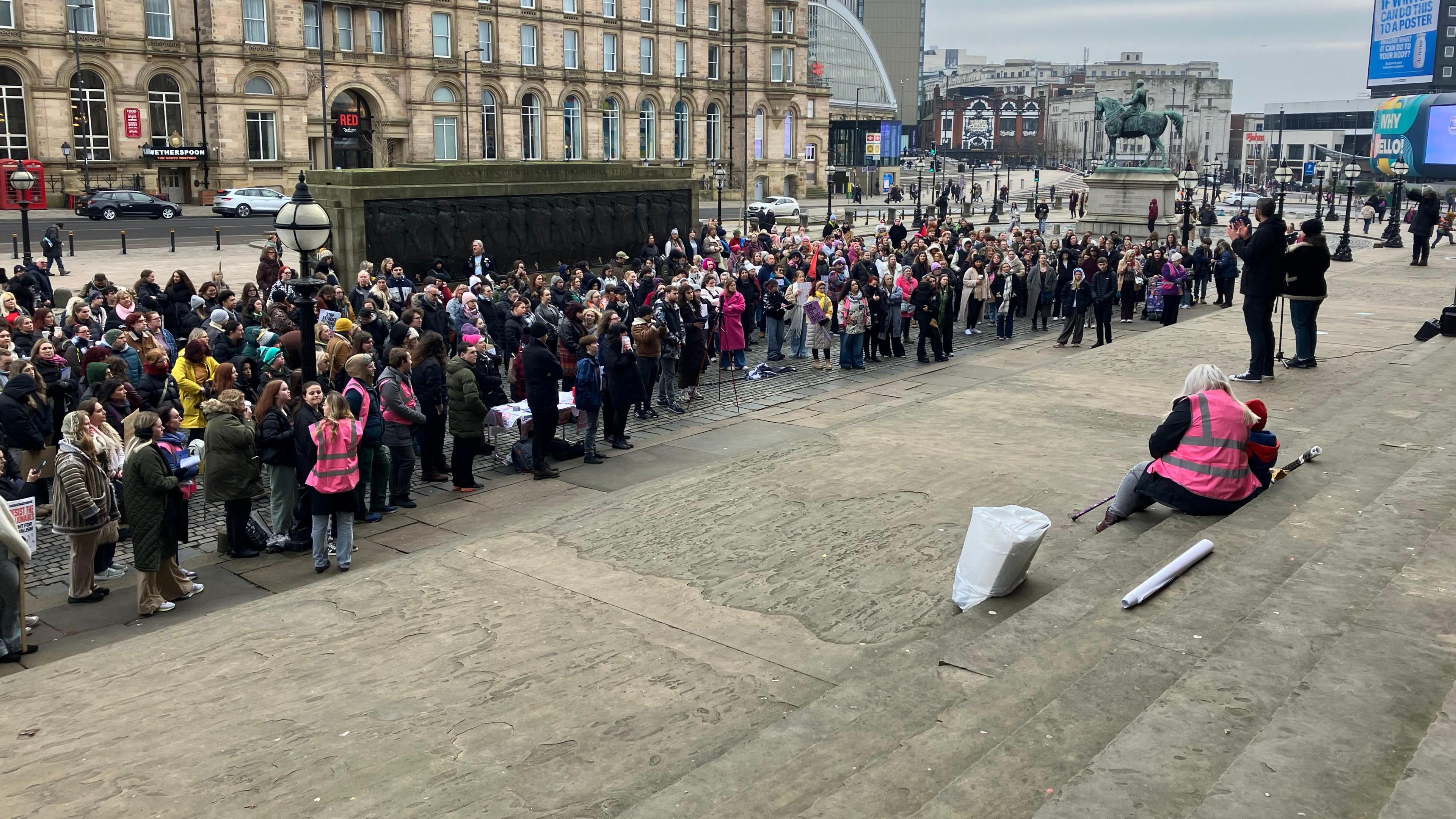 A crowd of people at the bottom of the steps of St George's Hall in Liverpool. Some are carrying placards and they are listening to a speaker on the steps.