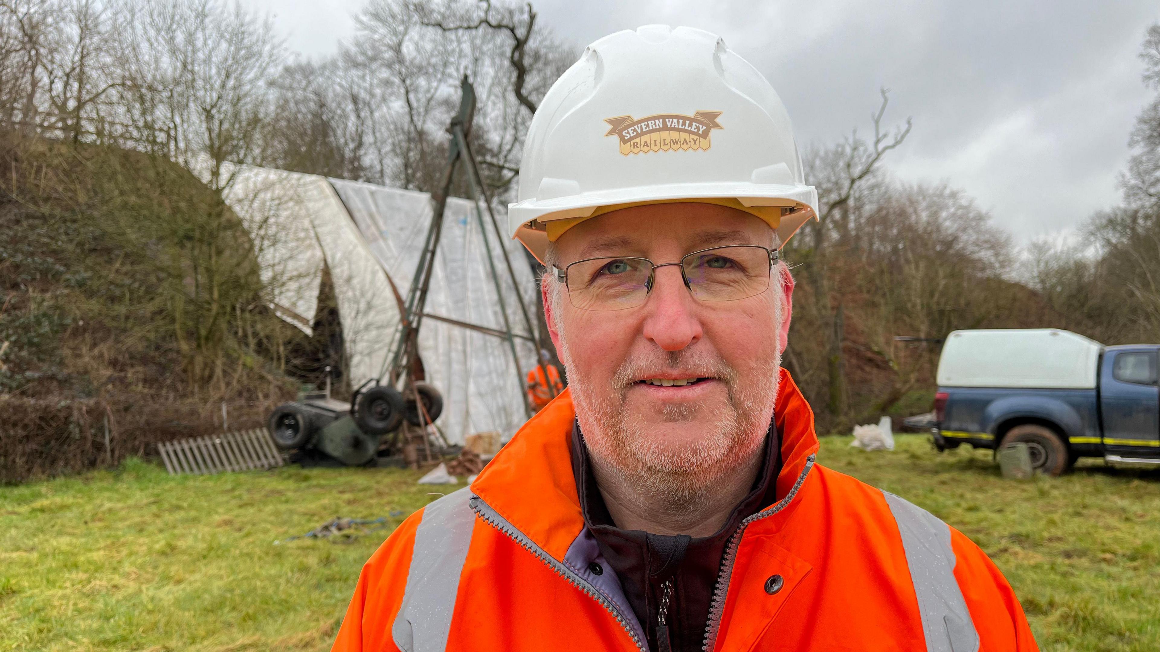 A man, Gus Dunster, is pictured in front of the percussive borehole - a huge, triangular metal structure. Beyond that, is a damaged bridge covered in white sheeting. He is wearing a white hard hat with a label that reads "Severn Valley Railway", and an orange high-visibility jacket. He is standing in a green field with trees and shrubbery on the edges. Behind him on the right is a truck. 