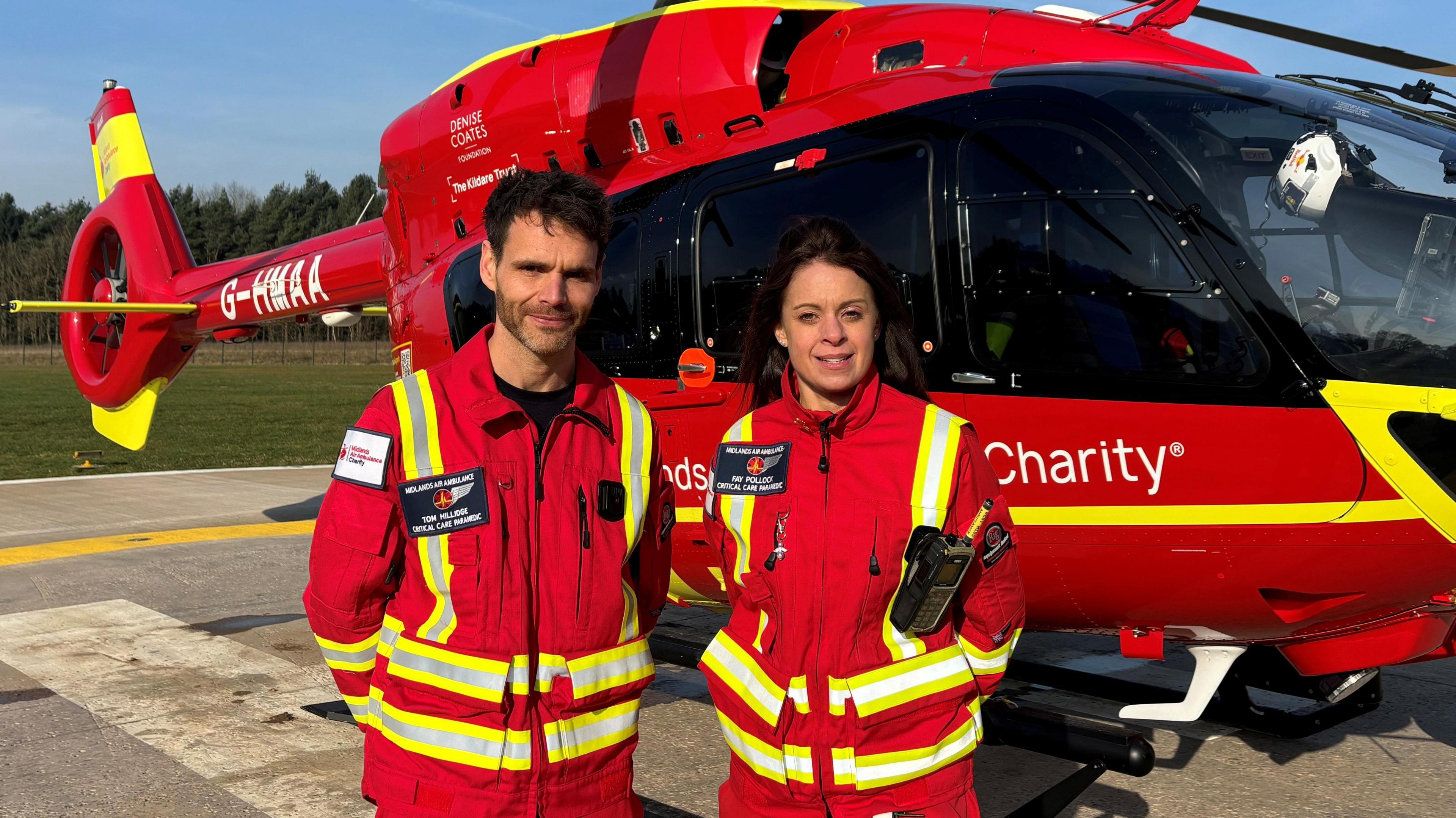 Two paramedics standing in front of helicopter with their hands behind their backs looking into the camera. 