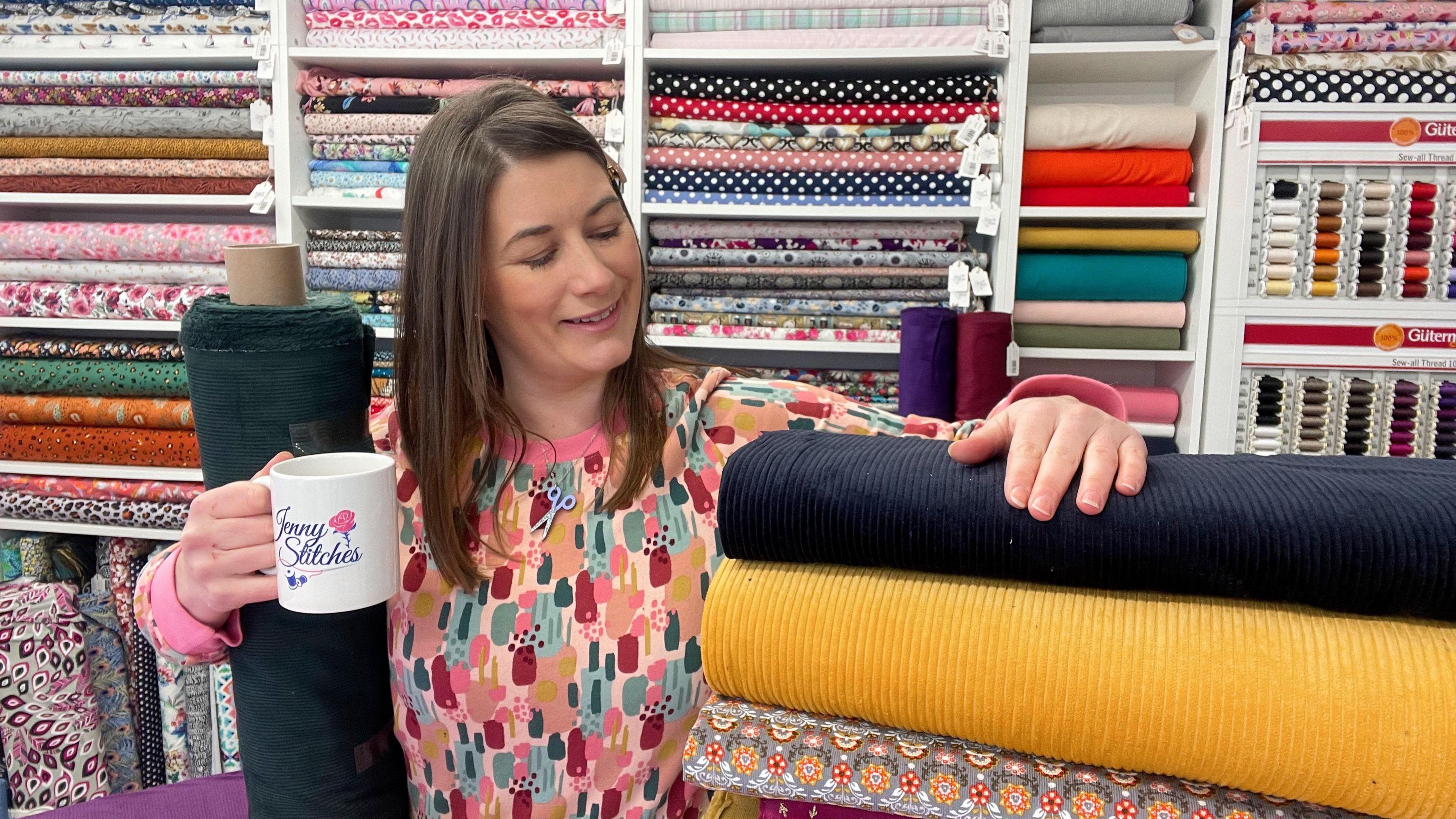  A medium close up of Jenny Fazackerley, owner of Jenny Stitches in Barrow, stood her shop, checking fabric, and holding a mug of tea.