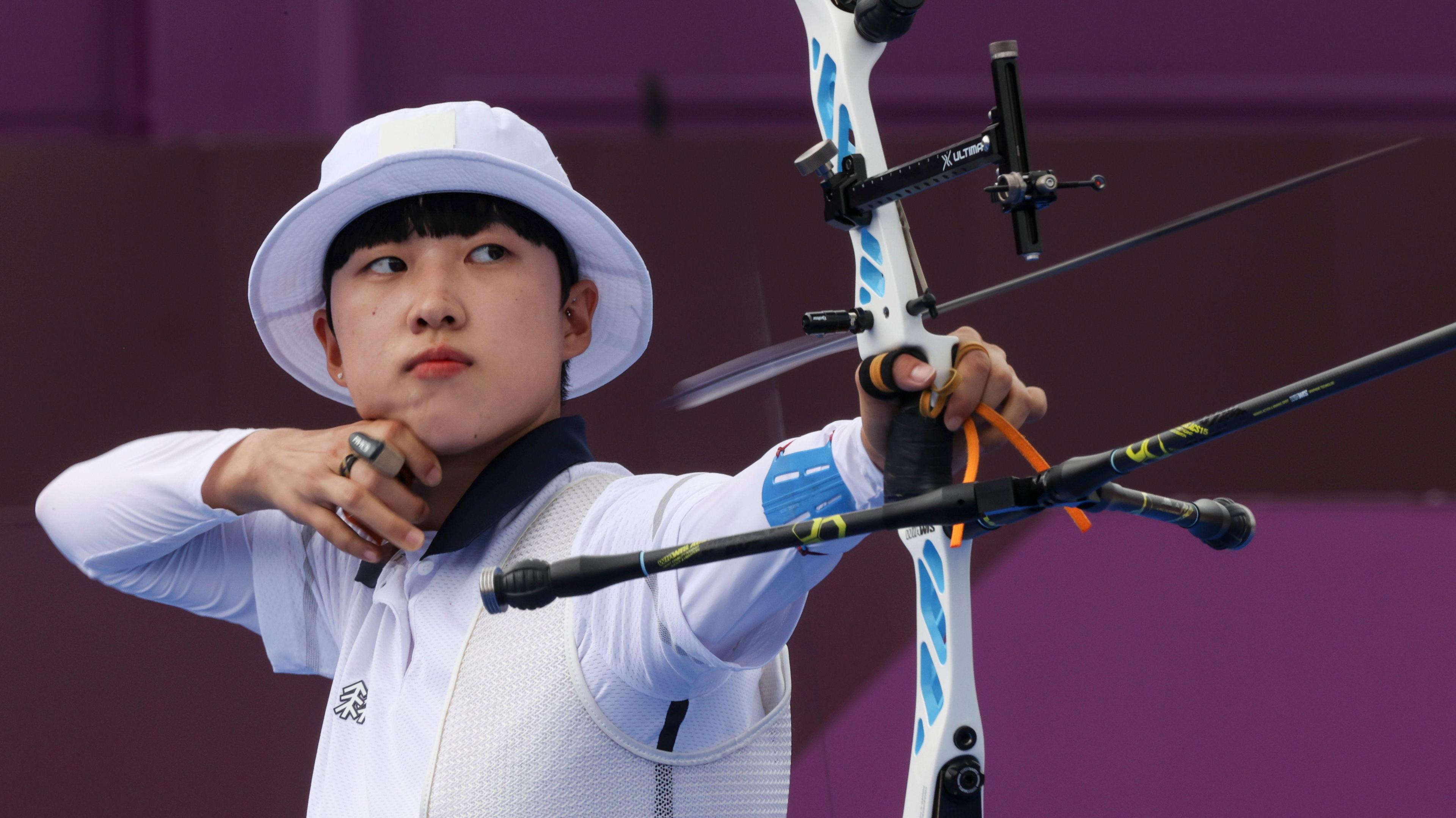 San An, wearing a white hat and long sleeved shirt fires an arrow. She is looking to the right of the camera