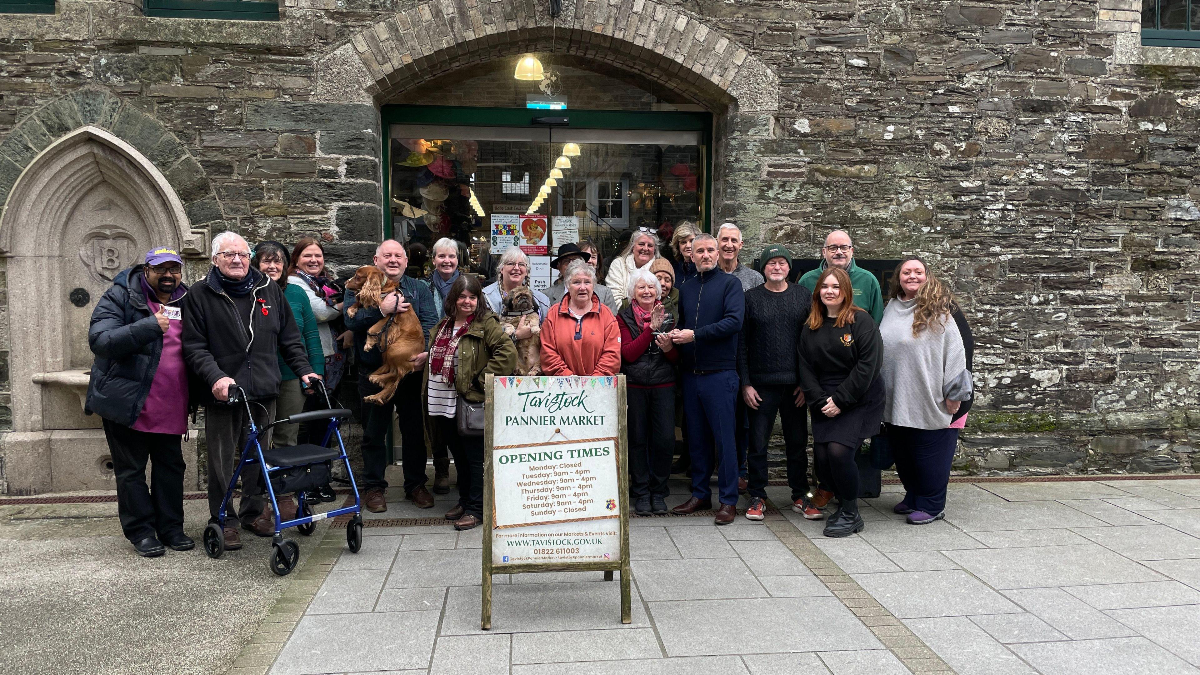 A group of traders gather outside of Tiverton Pannier Market. The group, made up of about 20 people, are standing in front of a stone wall. A sign which shows the market's opening hours is in front of the group. 