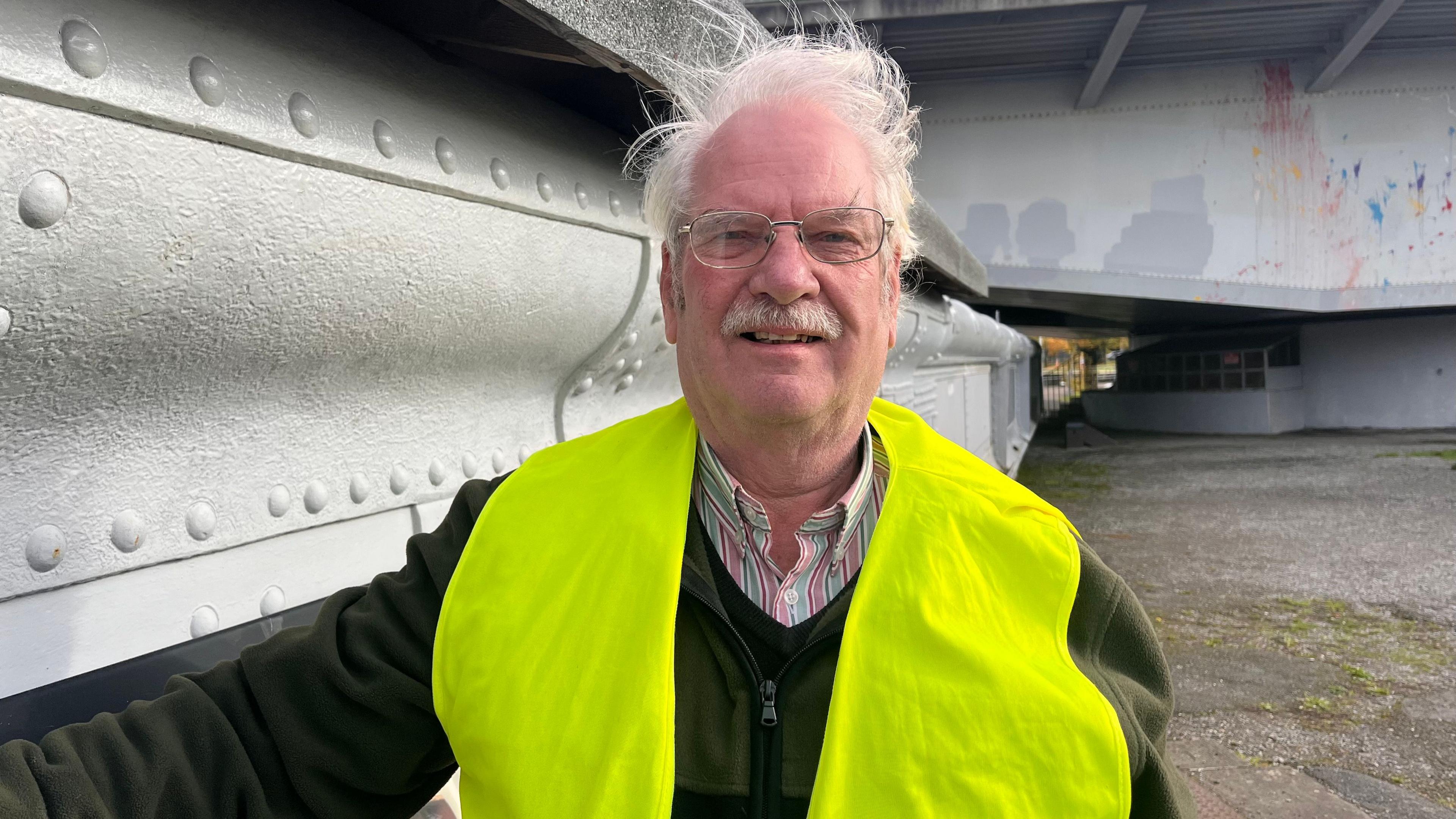 Wearing a high vis vest over a dark green jumper, Geoff Wallis looks at the camera, while stood next to the bridge. He wears oval glasses. 