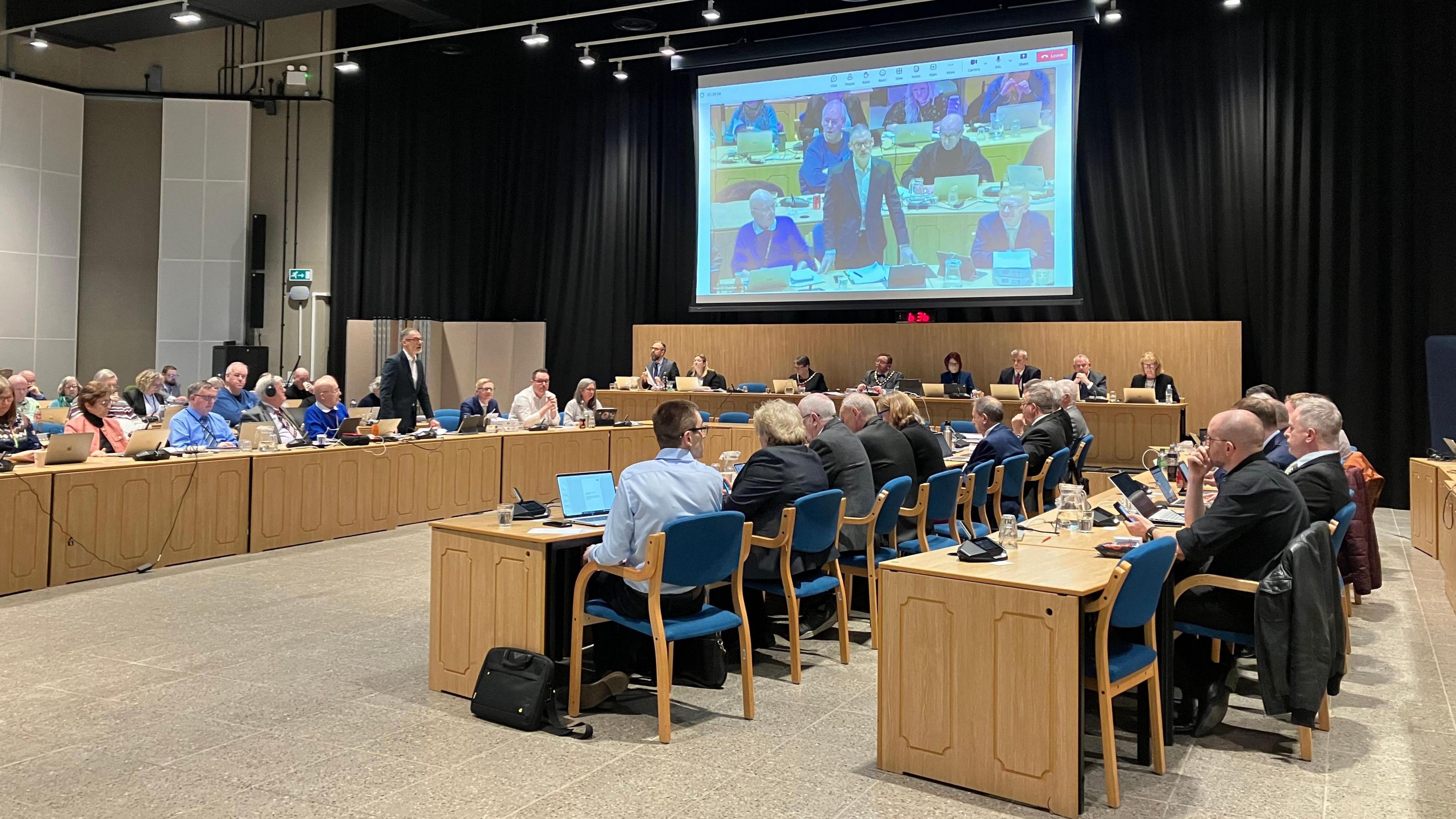 The council chamber with members sitting at five rows of light wooden desks and blue chairs, arranged in a U shape .  There is a screen above to "top table" showing the person speaking, which has black curtains pulled behind it. 