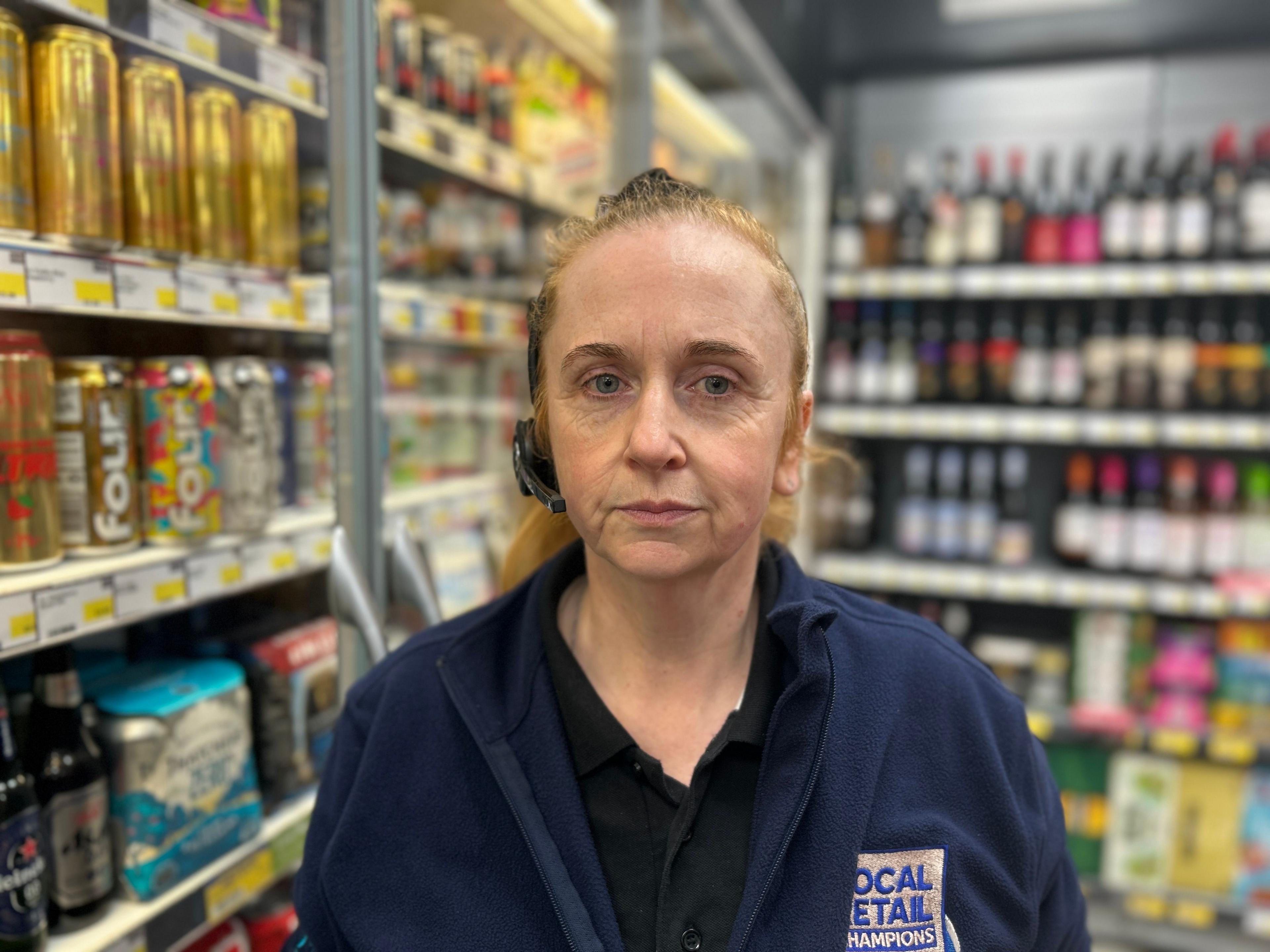 A woman wearing a work uniform and a headset stands in the aisle of the shop that she works in. She is standing in front of a fridge with drinks inside.
