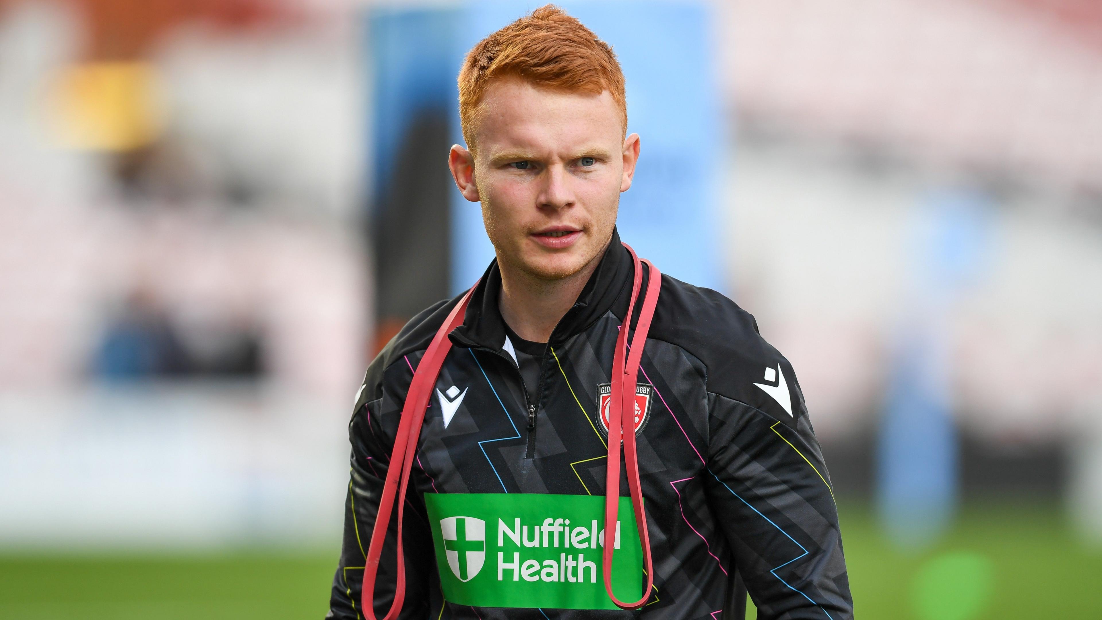 Scrum-half Caolan Englefield warming up for a Premiership Rugby match with Gloucester, wearing a black training top, with red stretch bands around his neck. 
