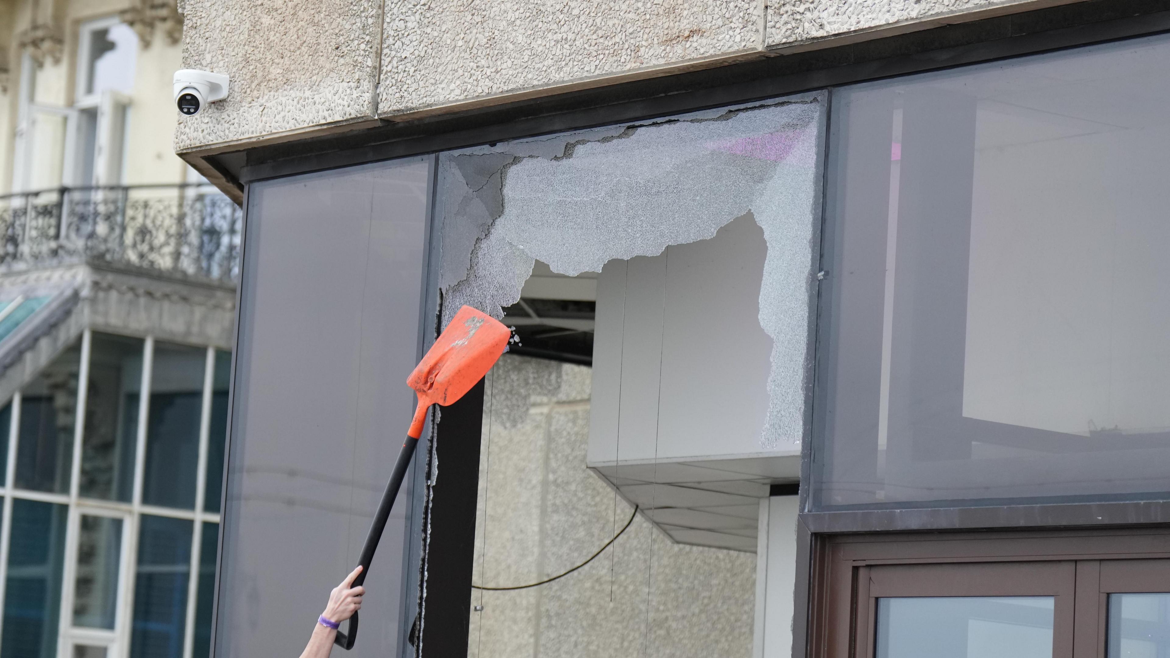 A window pane on a building is smashed. Someone is poking the broken glass with a spade.
