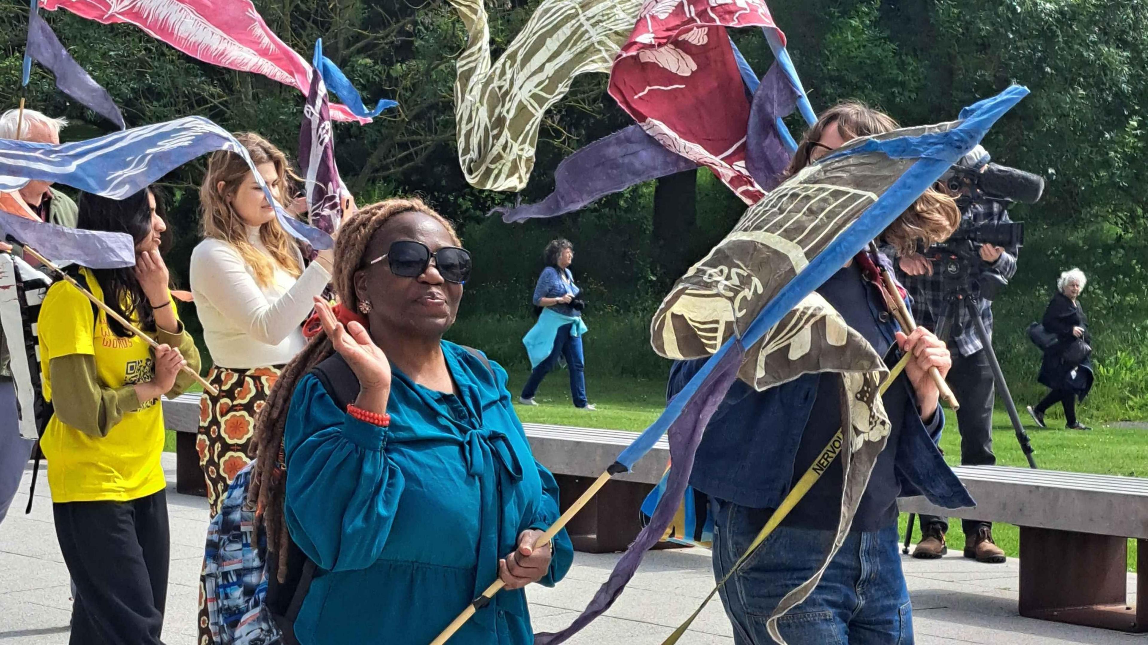 People are holding flags and banners on wooden polls on a concrete walkway. The flags are all different patterns and are held in the air. In the background are a row of benches and trees.