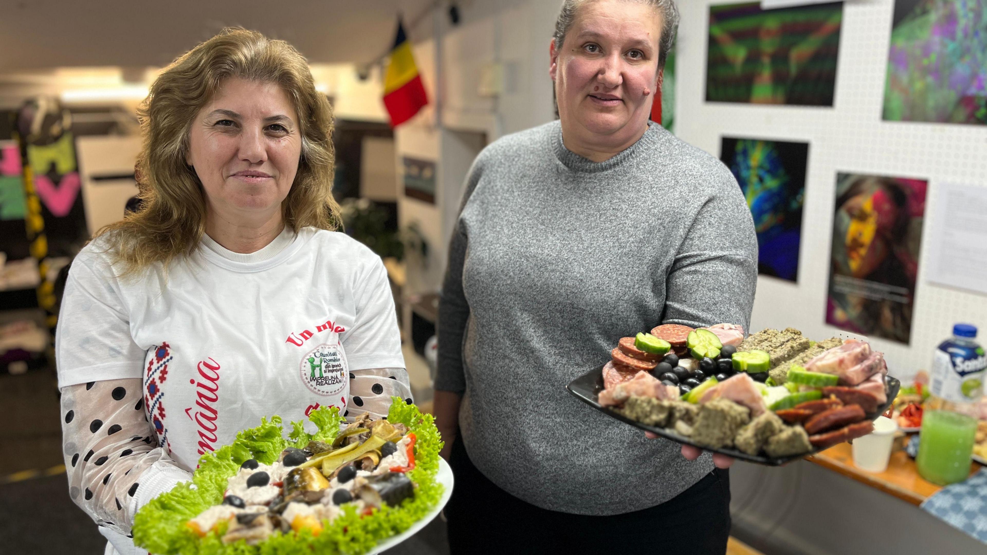 Two women hold plates of food, with fish, salad and meat products. They are standing inside. On the left, the woman is wearing a white t-shirt. On the right, the woman is wearing a grey round-neck jumper.