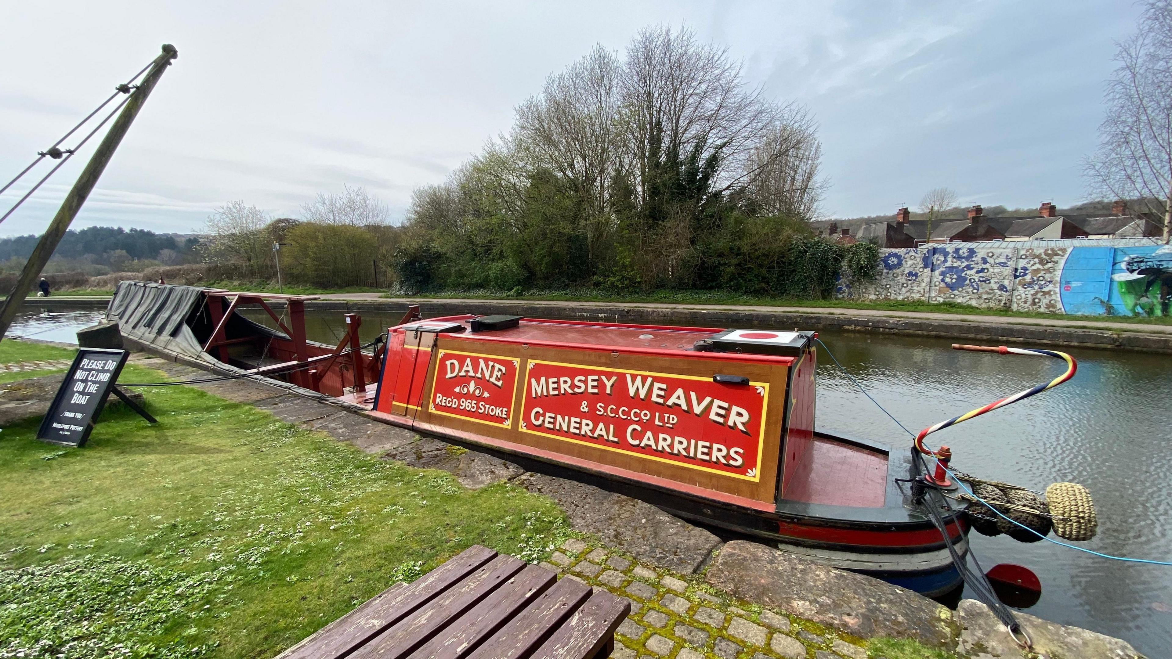 A red narrowboat with writing on it reading "Dane" and "Mersey Weaver and S.C.C.Co Ltd General Carriers" in a canal. A picnic table, a black sign and grass are on one side of the canal while a wall and trees are on the other side.